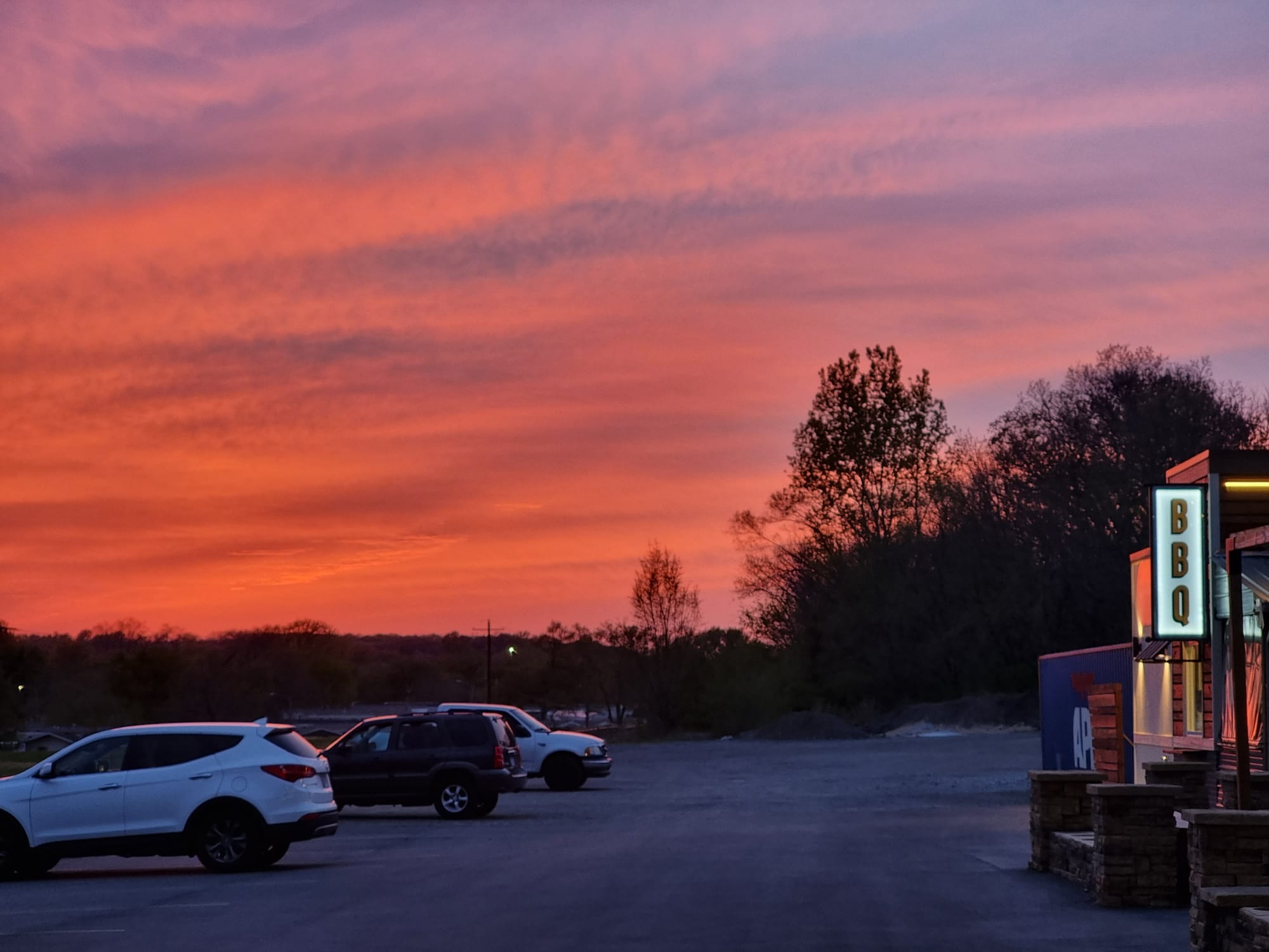 The Midwest sun in the background - a pinkish hue. In the foreground a parking lot with a few cars and to the side the steps of a restaurant and a sign saying "BBQ".