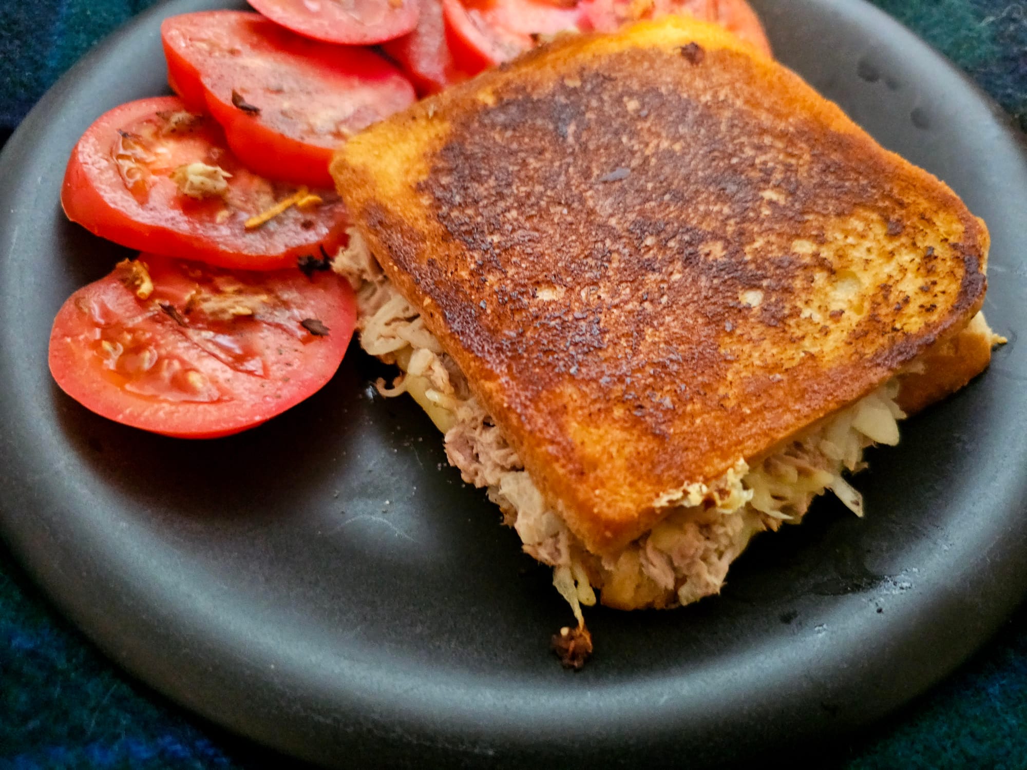 A tuna melt on a black plate with a cut up tomato as garnish