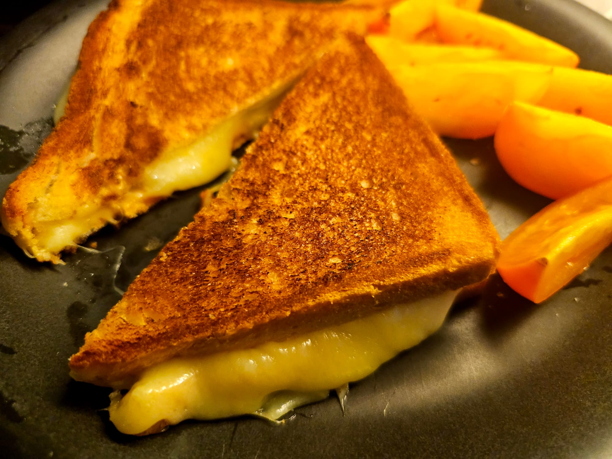 A grilled cheese sandwich, cut in two triangular pieces, cheese oozing out at the sides. Behind it a cut up yellow tomato. 