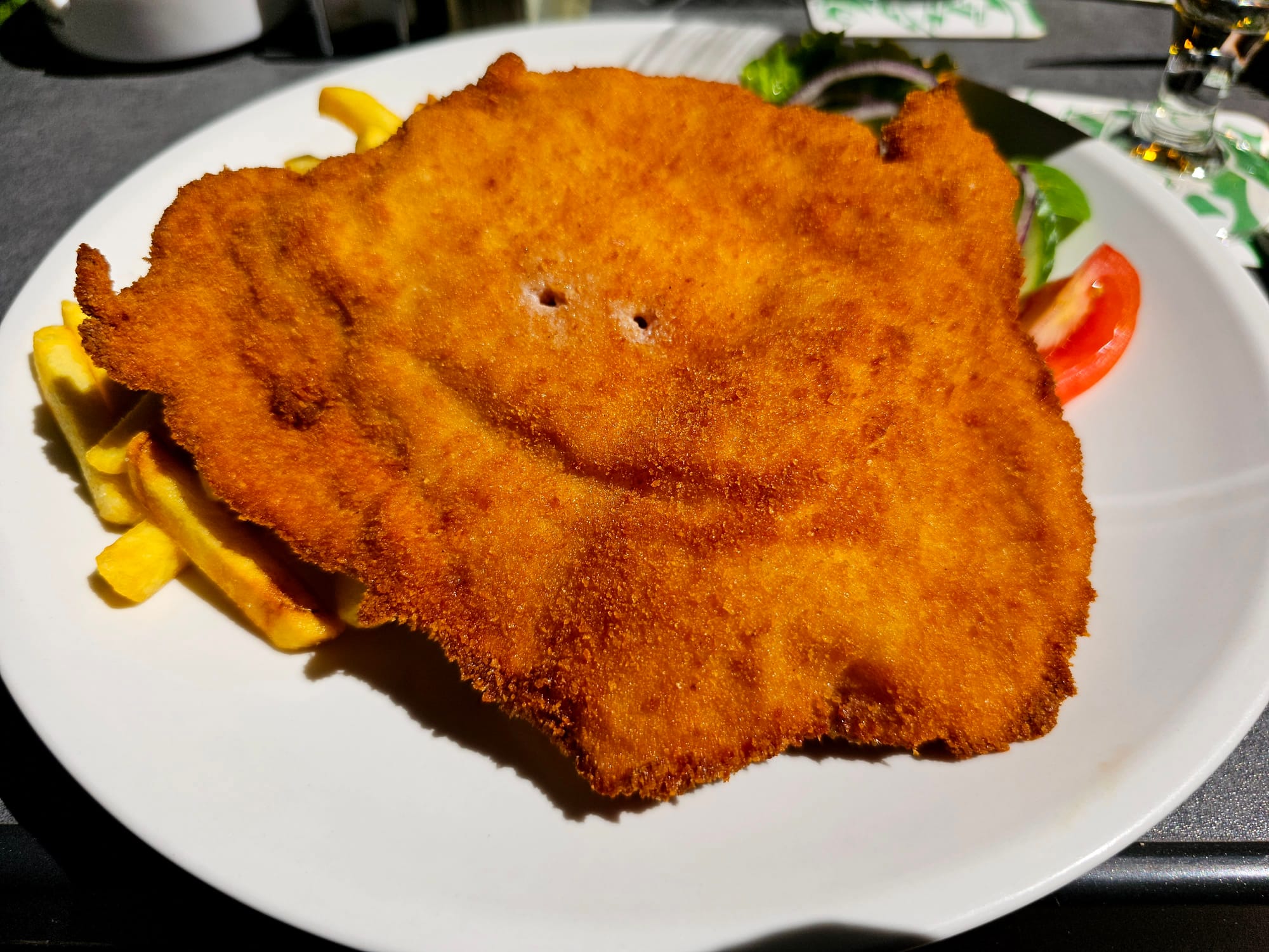Image of a cordon bleu on a bed of chips, on a white plate. Two fork holes, in combination with a fold in the batter of the cordon bleu give the impression of a sly smile. 