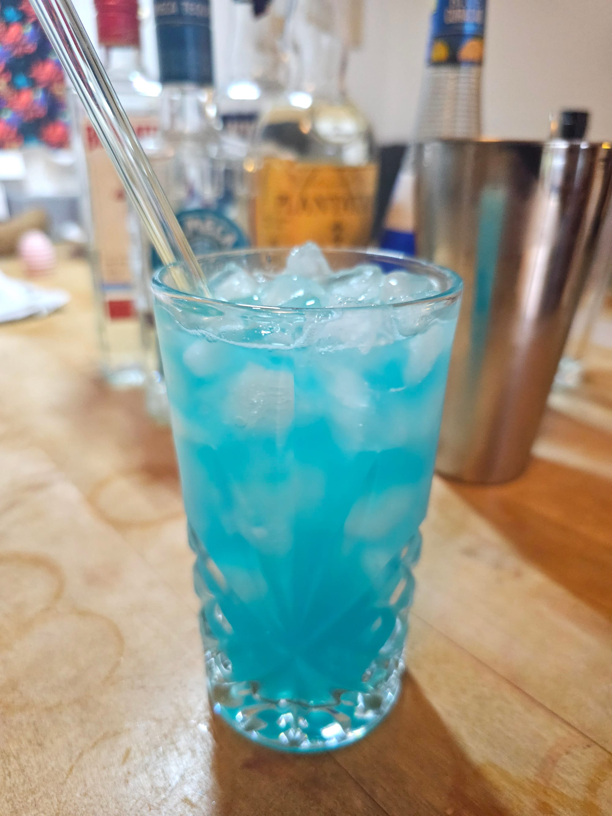 A longdrink glass filled with a blueish liquid and ice. In the background cocktail paraphernalia and bottles. 