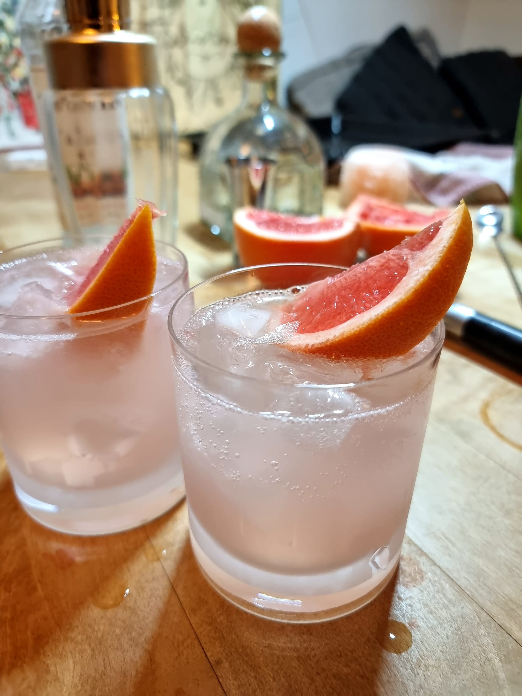 Two double old-fashioned glasses, filled with a pink, slightly sparkling liquid and some ice. Garnished with a slice of grapefruit. In the back, out of focus, a few bottles and cocktail paraphernalia.