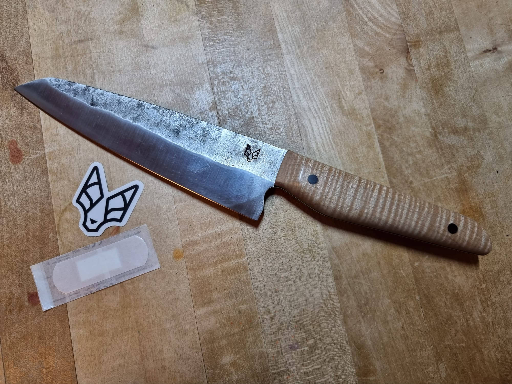 A kitchen-knife with a wooden handle on a wooden table. Next to it a sticker with maker's logo (a stylised fox?) and a band-aid.