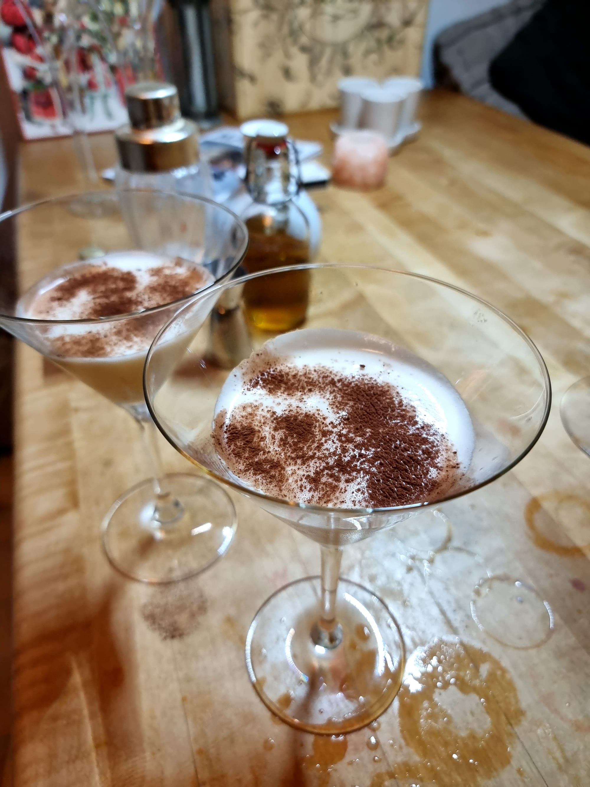 Two Martini glasses, from above. White liquid, powdered brown cocoa powder on top. On a wooden table that's showing signs of many previous cocktails.