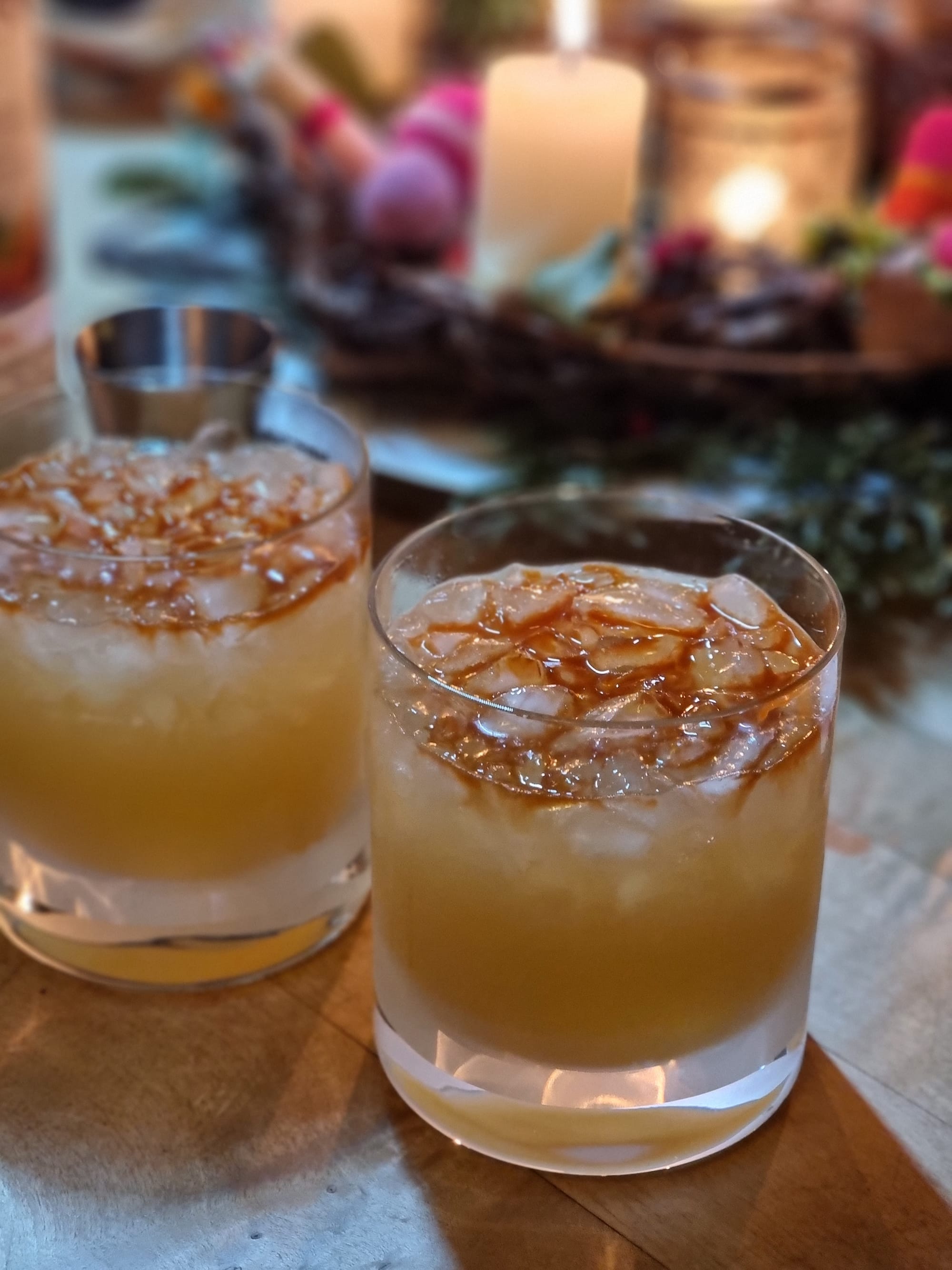 Two double old fashioned glasses filled with a yellowish liquid and crushed ice. On top the magical pattern of five dashes of reddish brown Angostura bitters spread out among the little pebbles of crushed ice. In the background, blurry, a wreath with burning candles.