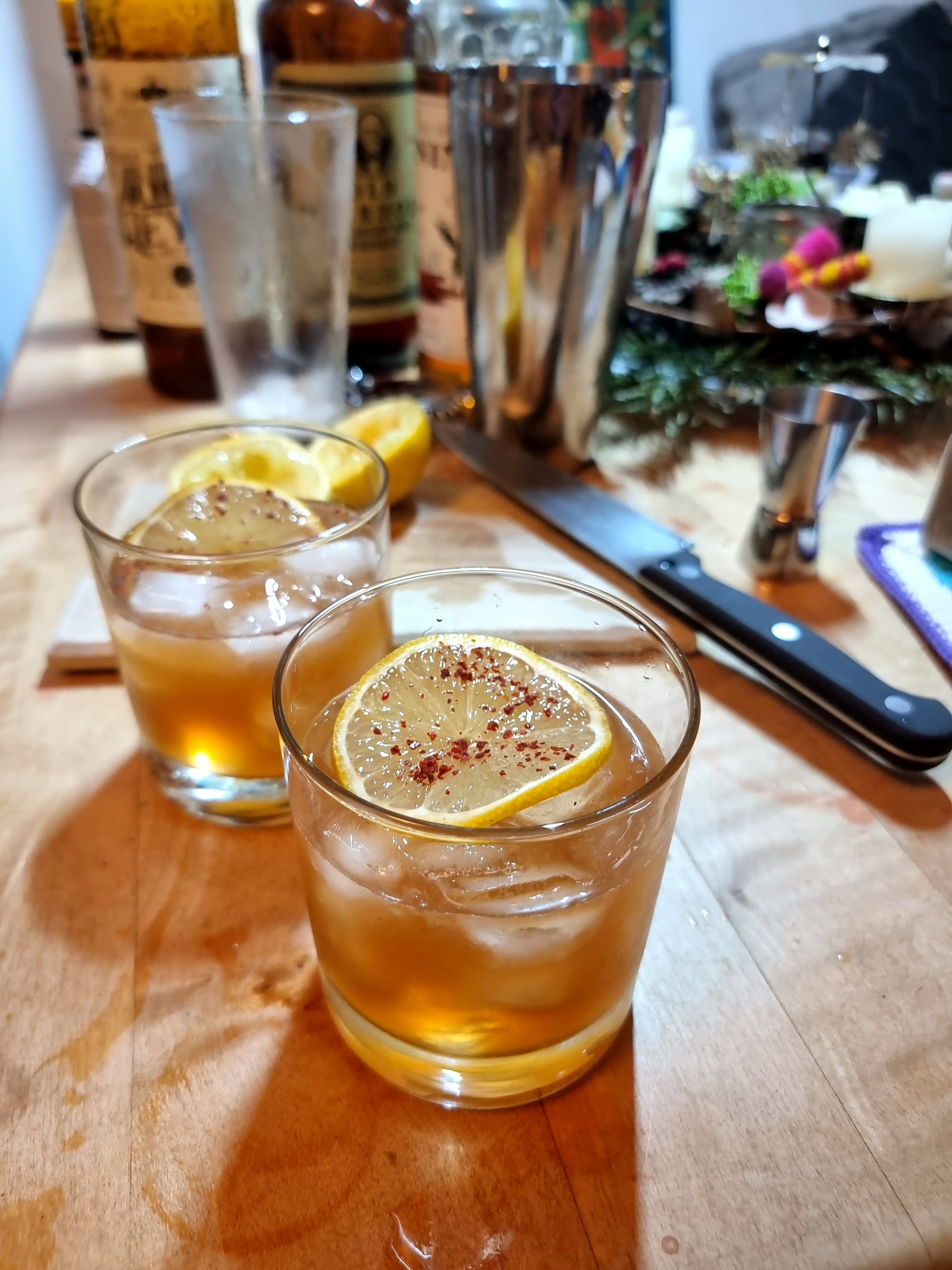 Two double old fashioned glasses, filled with a golden liquid and ice, garnished with a slice of lemon and chili flakes. In the background the usual cocktail paraphernalia.