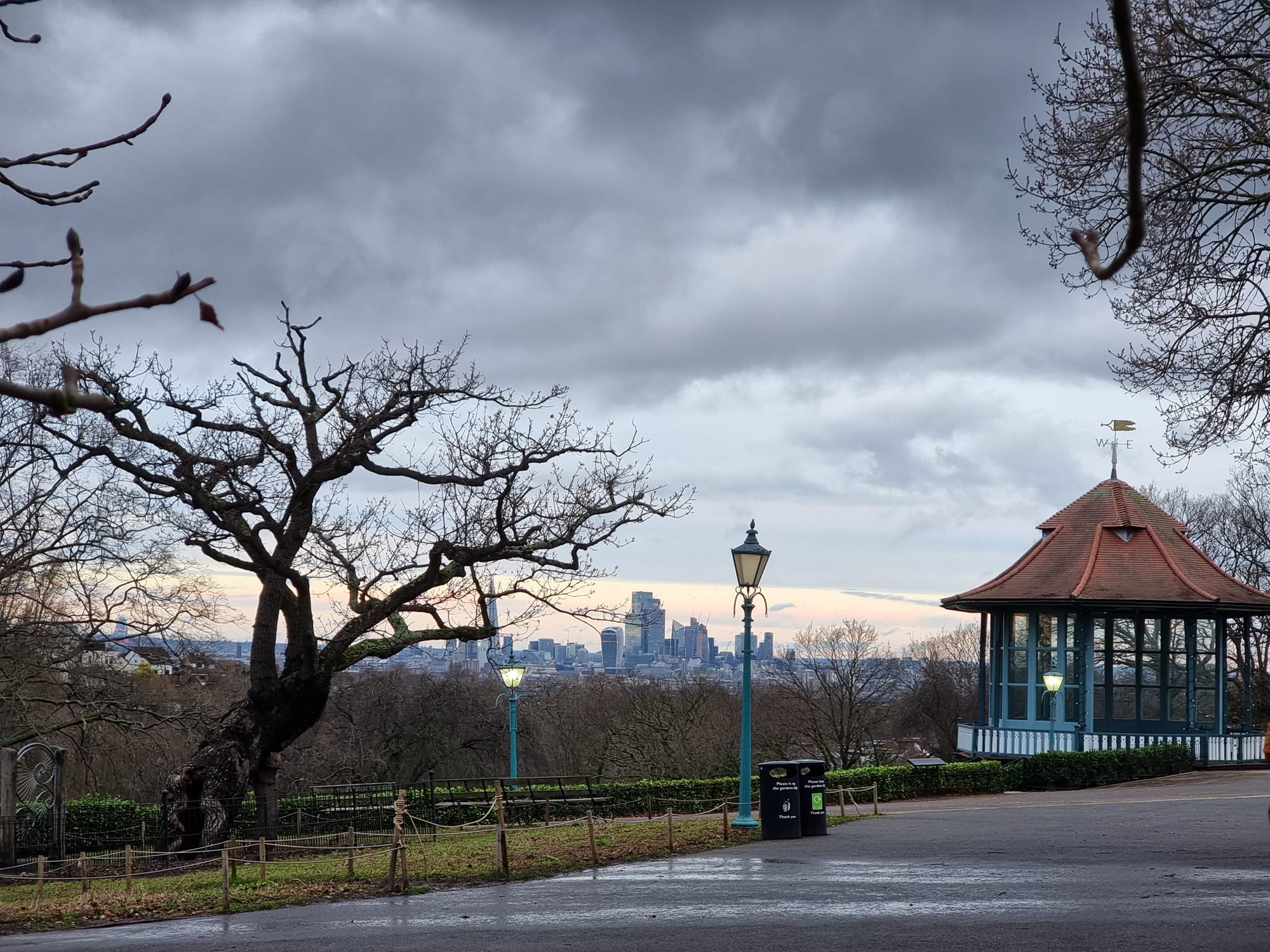 View into the skyline of Central London from a hill in the South. A gnarly tree to the left, an arbour to the right, a street-lamp in the centre and a grey-blue sky above.