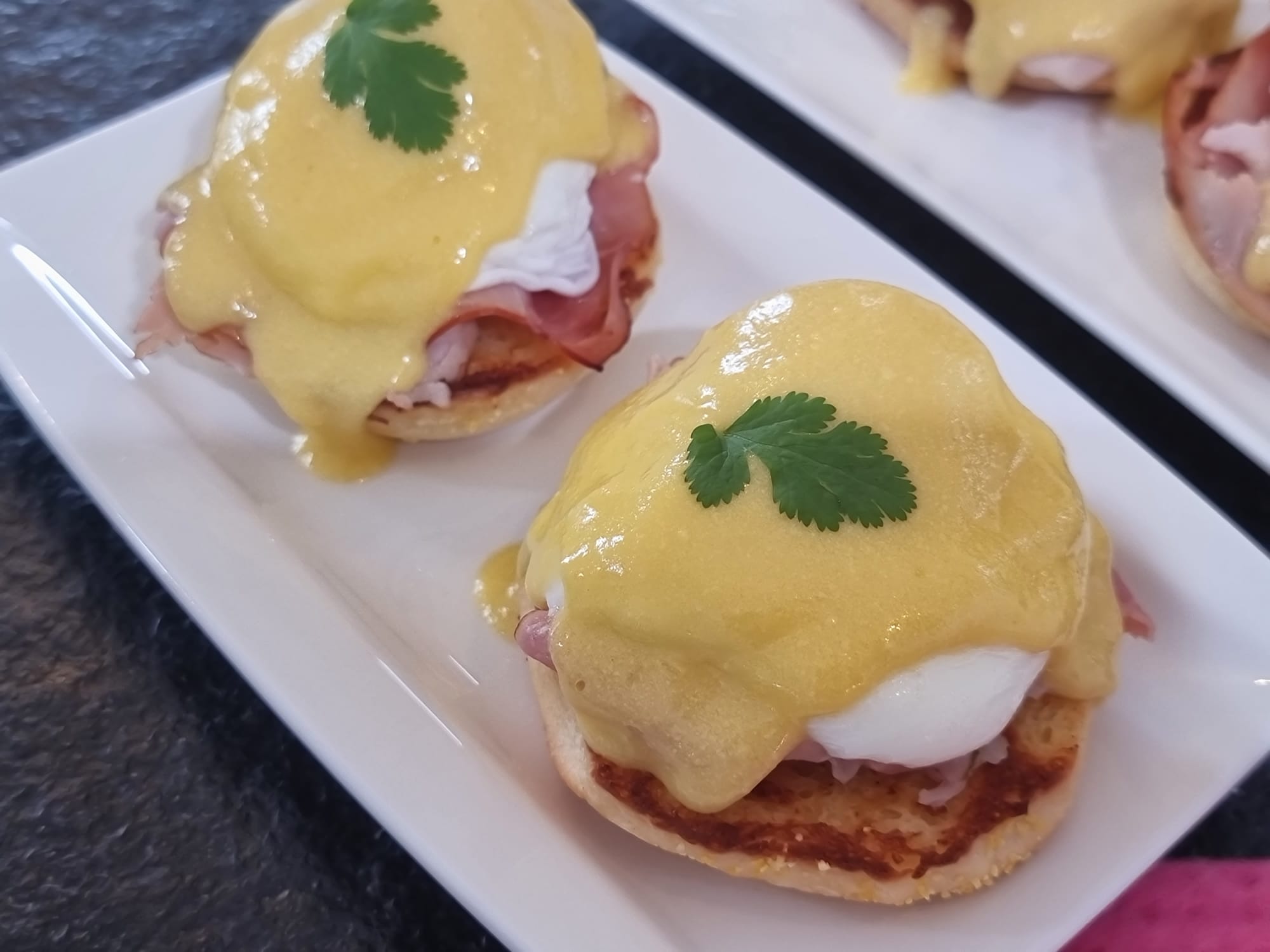 Two Eggs Benedict on a rectangular white plate. Grilled English muffin, grilled ham, poached egg, covered by a thick Sauce Hollandaise.