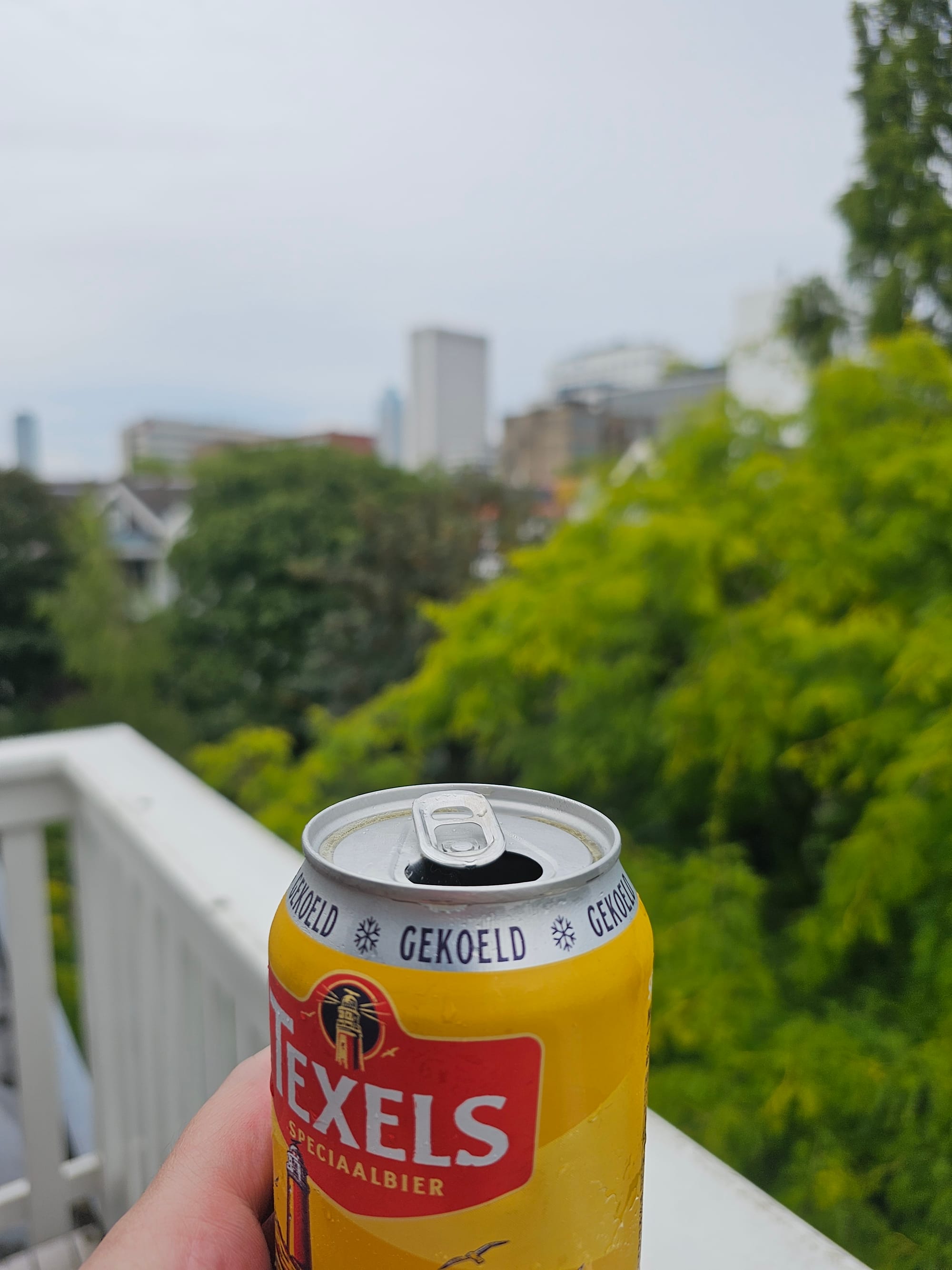 Can of Texels Speciaalbier in the foreground, a white balcony banister, lush greenery and some houses in the background 