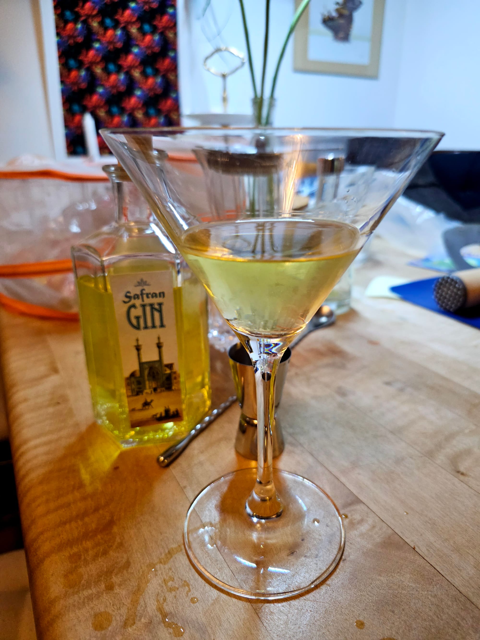 A martini glass, filled with a yellowish liquid. In the background a bottle of Saffron gin. 