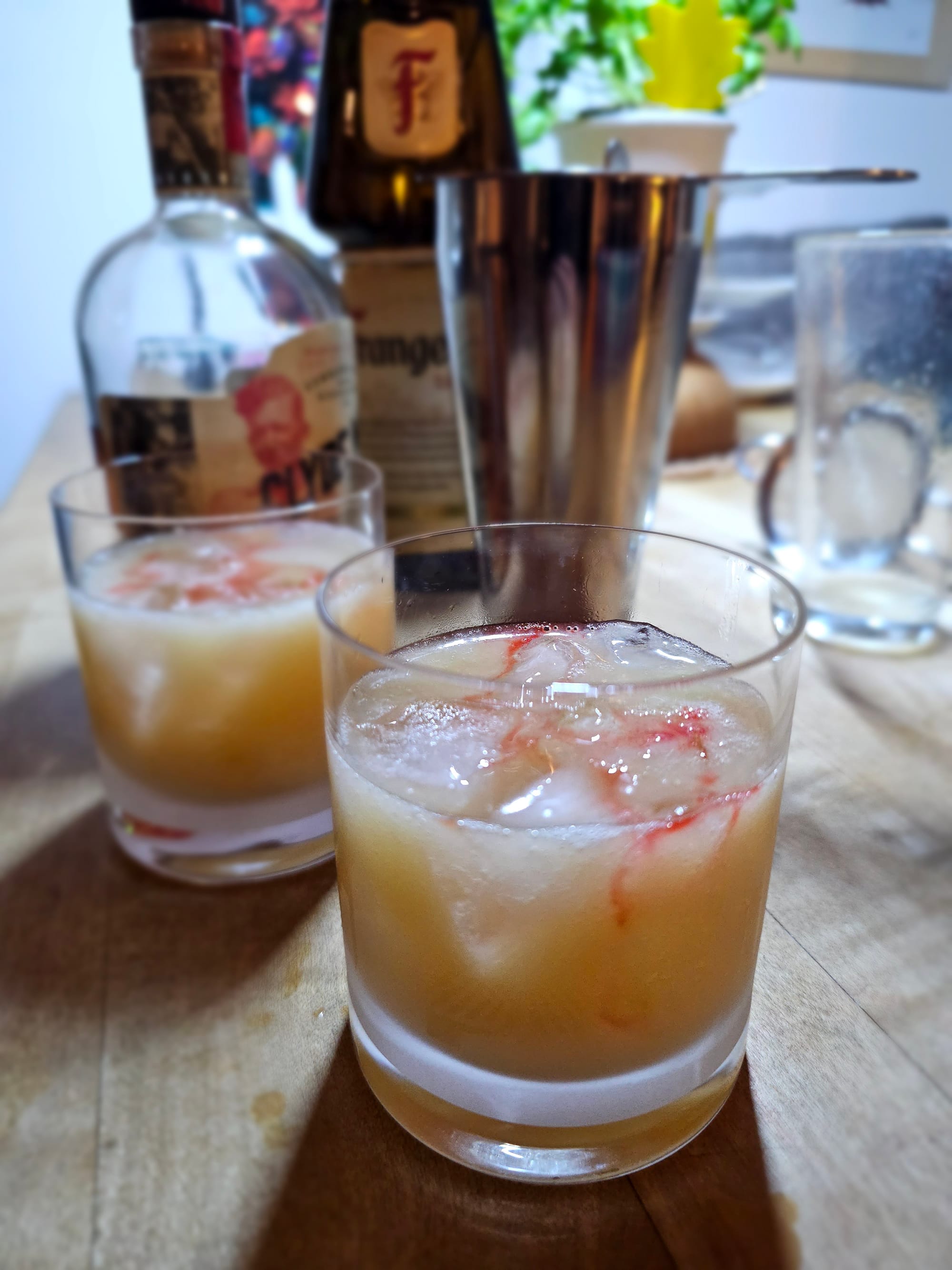 Two double old fashioned glasses filled with a milky, yellowish liquid and ice, red rivulets on top. In the background the usual cocktail paraphernalia. 