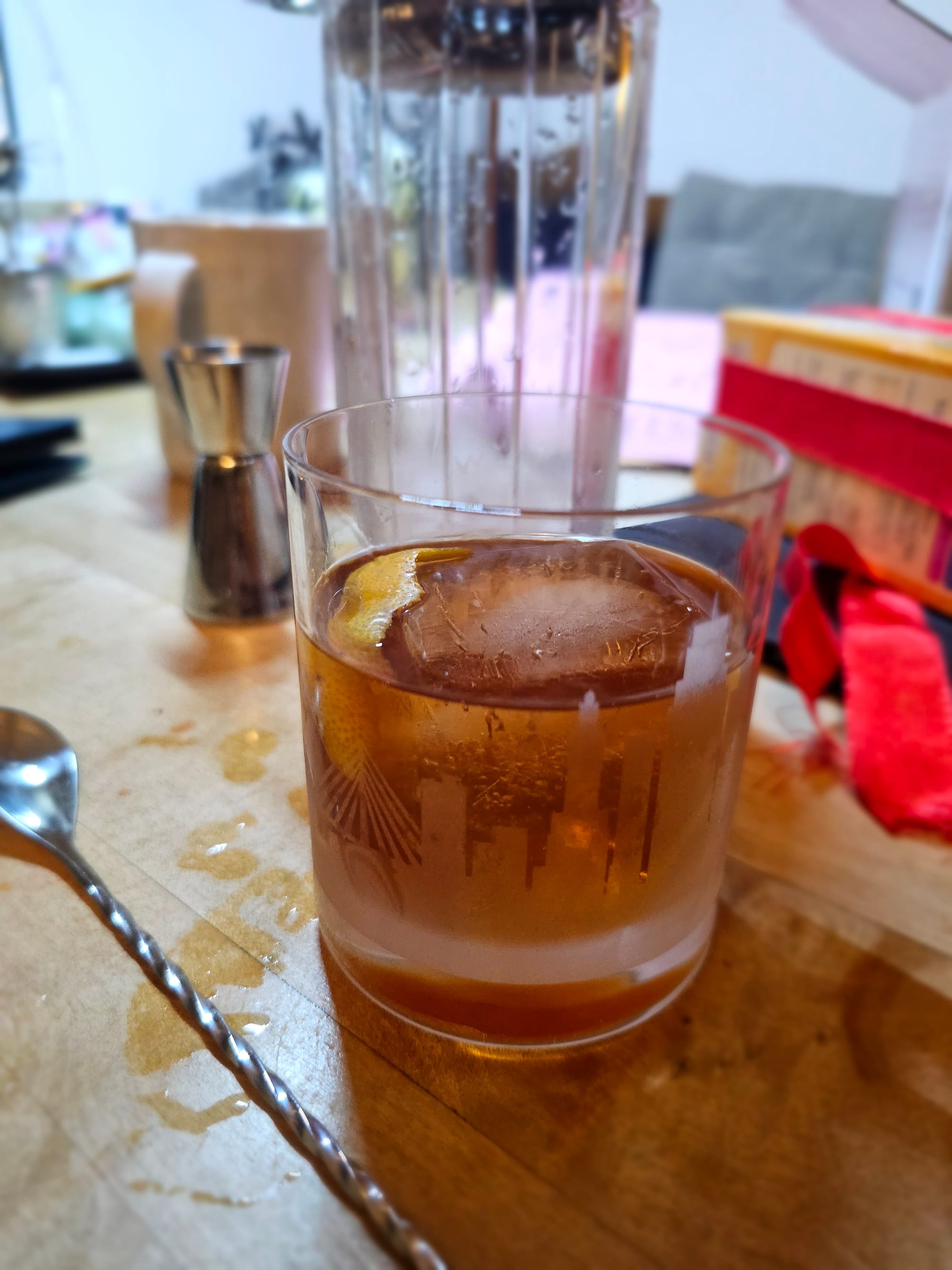 A double old fashioned glass filled with a big ice cube and a brown liquid. Next to it a mixing spoon, in the background a mixing glass and jigger. 