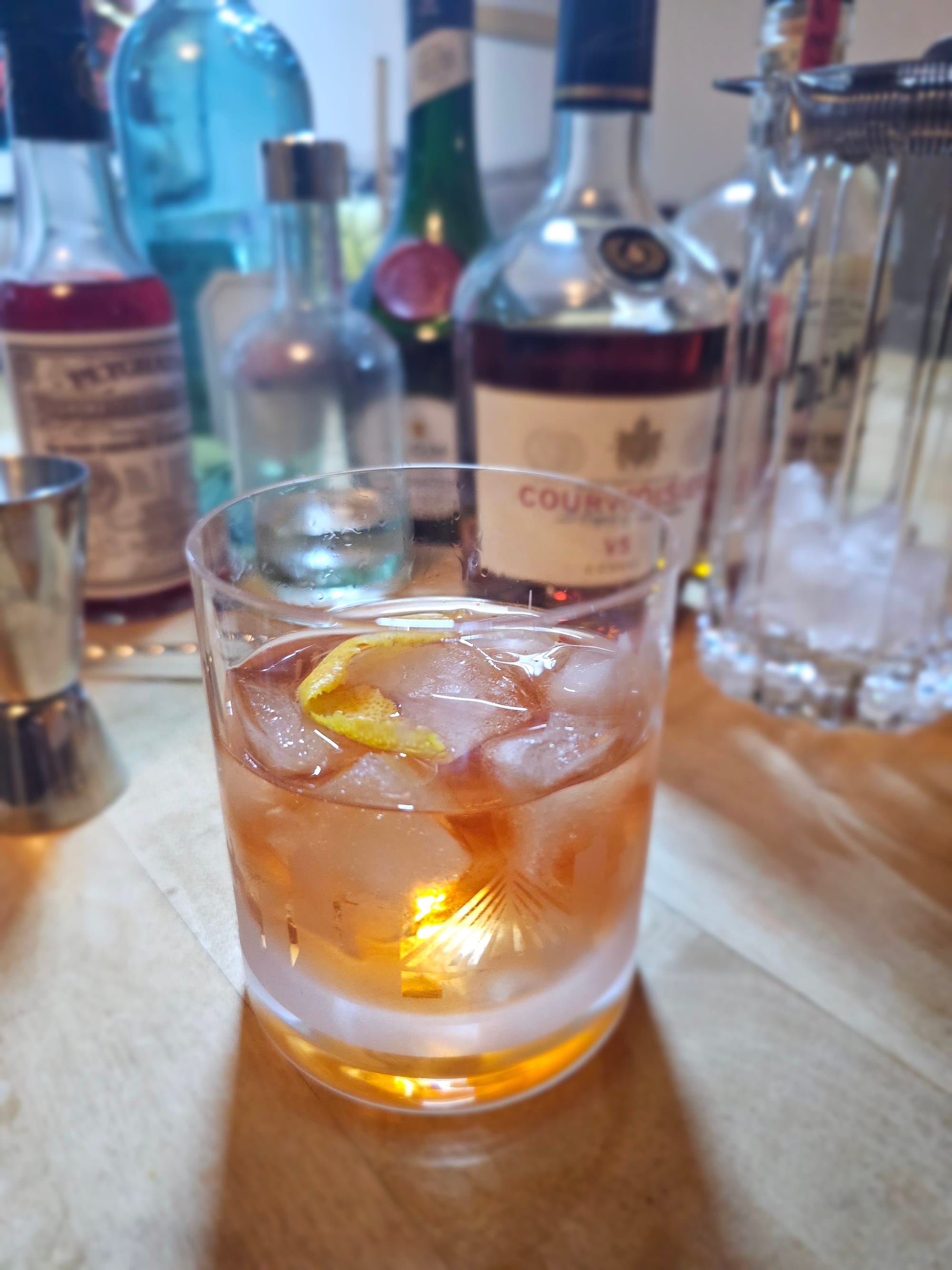 Double old fashioned glass filled with yellowish liquid and ice. In the background various bottles and cocktail paraphernalia 