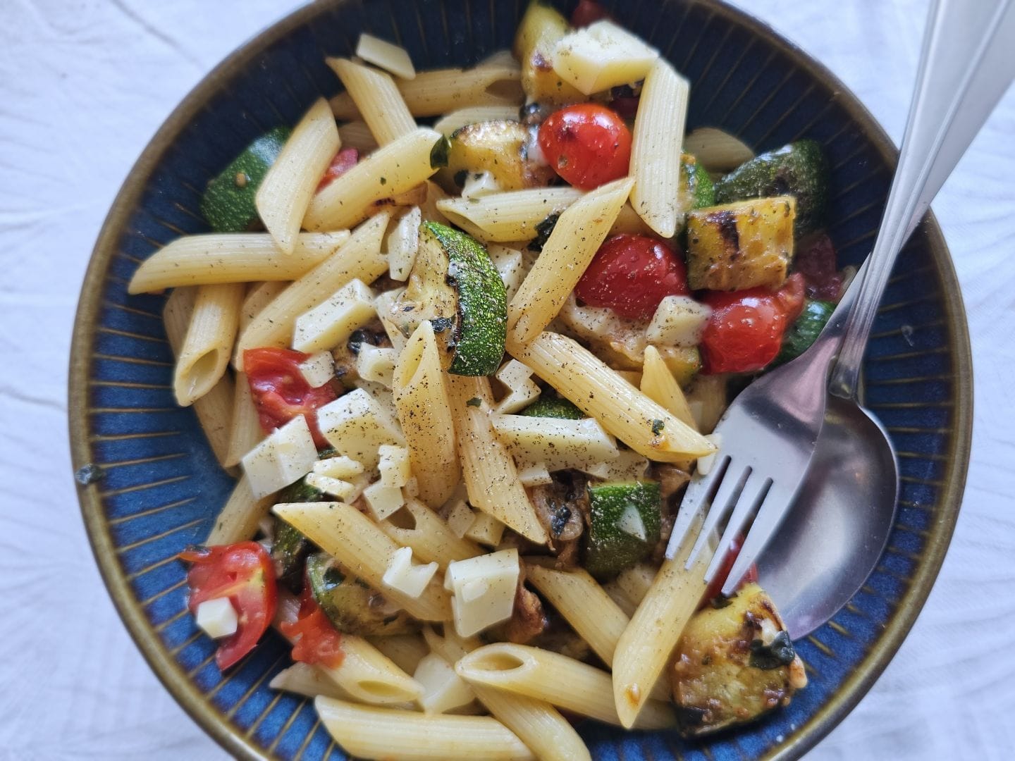 A blue plate of penne with bits of zucchini, cherry tomatoes and cheese