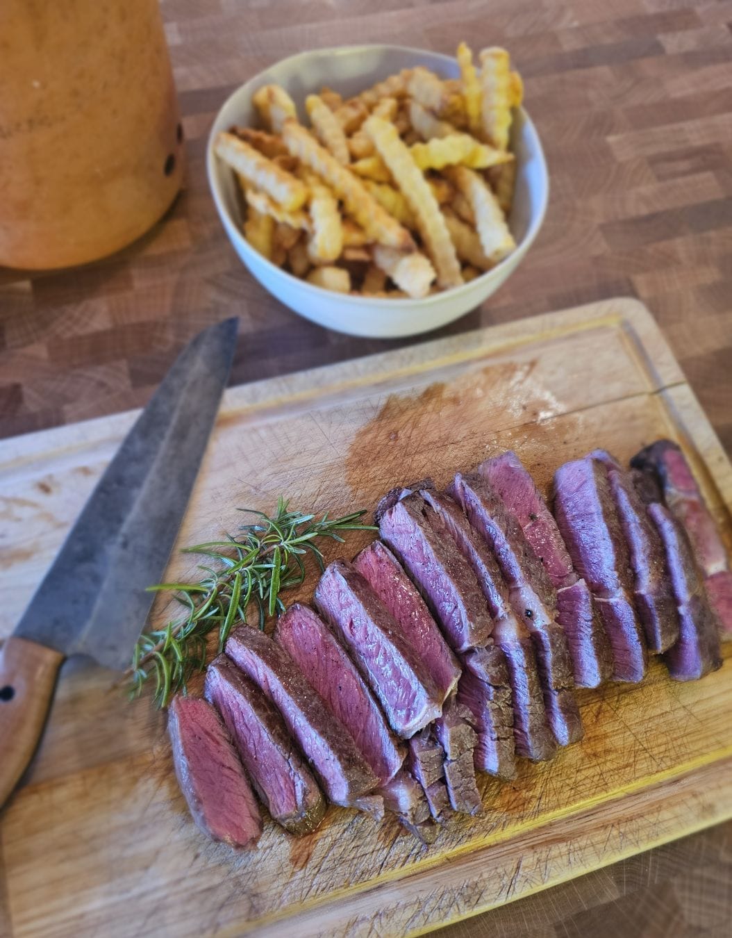 Steak cut up into strips, on a wooden cutting board. Some rosemary, a knife. In the background chips (or fries, depending on where you live).