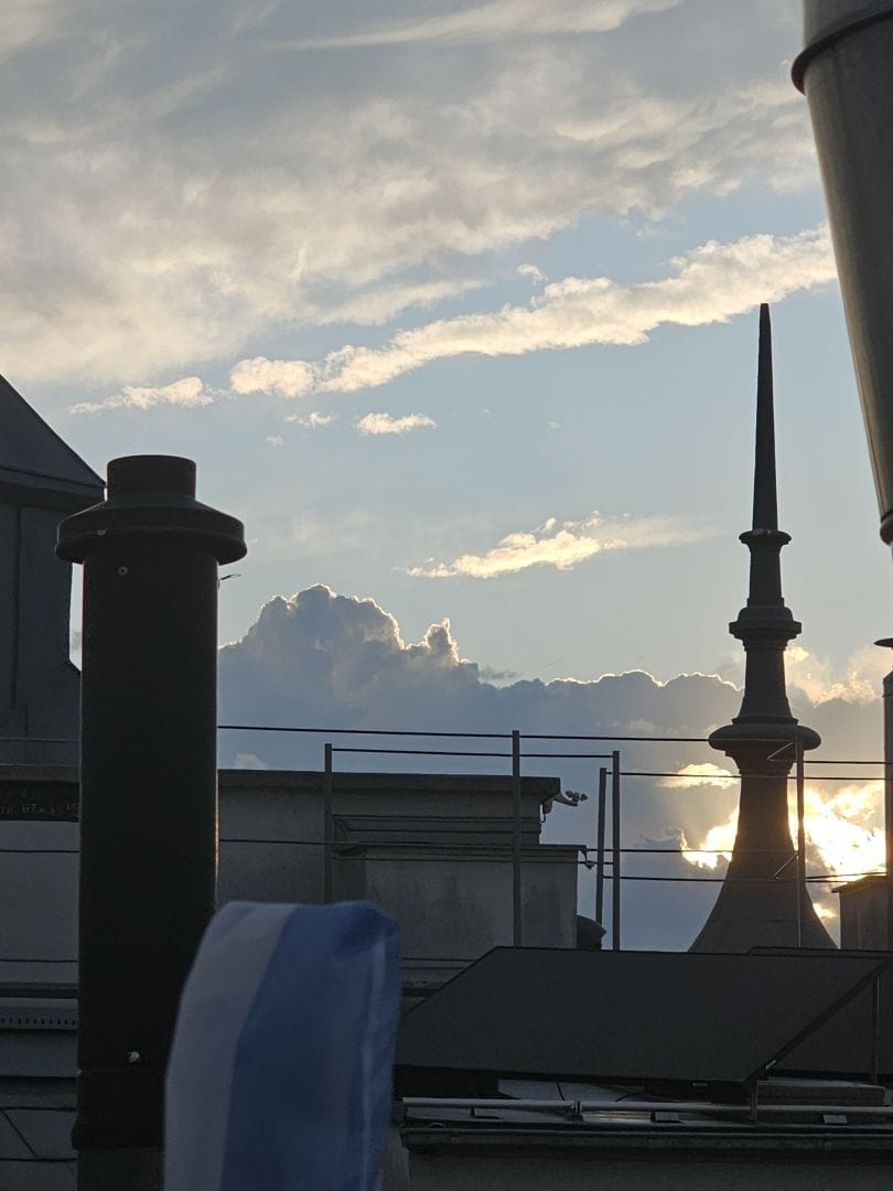 A cloudy blue sky with a setting sun, in the foreground various little rooftop chimneys and towers