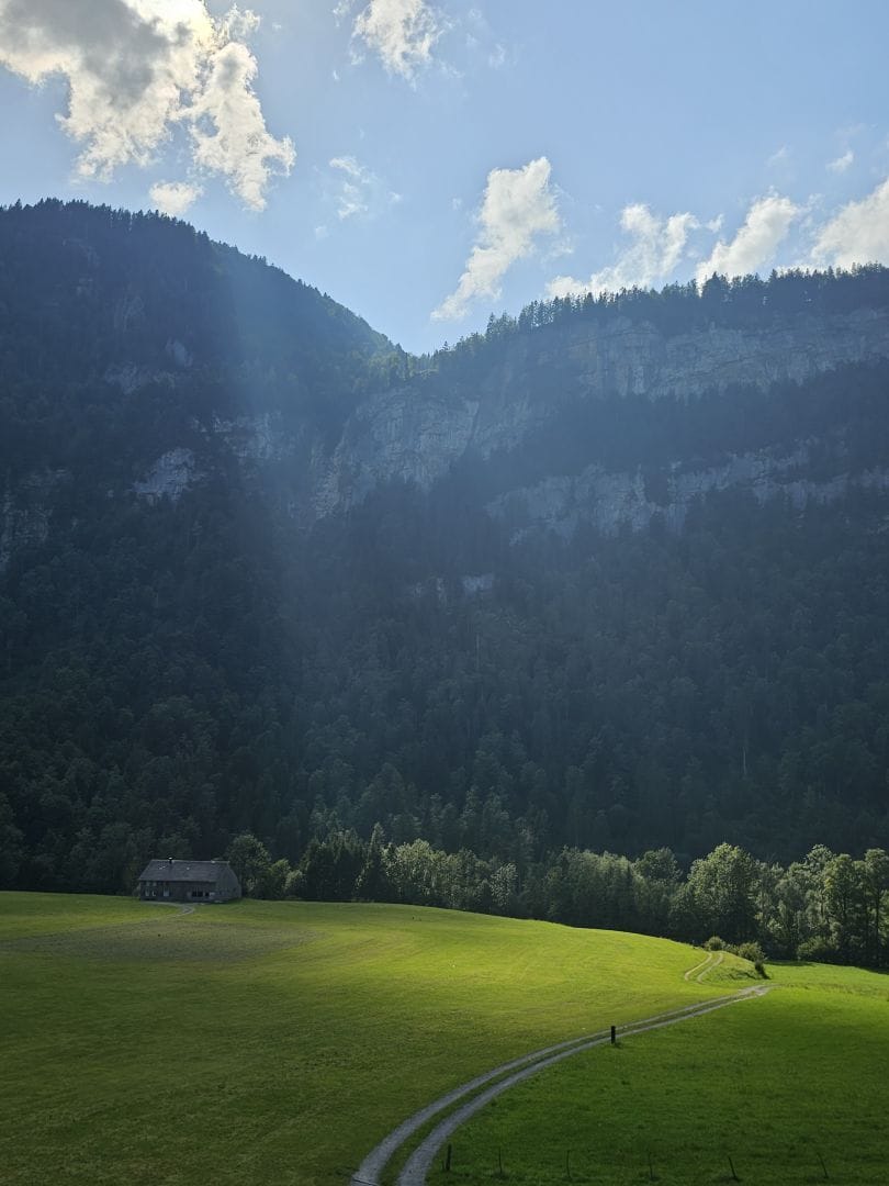 A meadow, partly illuminated by a beam of sunshine. In the background a farmhouse and imposing mountains. Also blue skies.
