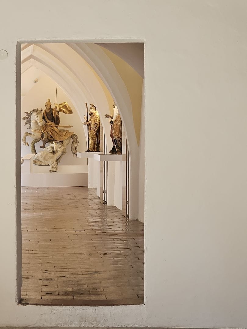 View through a doorway to a gallery with medieval-style statues, including a mounted figure on horseback and standing saints.