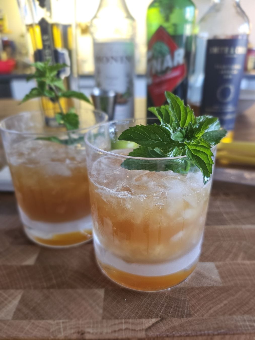 Two Two cocktails in short glasses filled with crushed ice, showing layered shades of amber, golden brown, and pale white at the bottom. Each drink is garnished with a fresh green mint sprig, sitting on a wooden surface with blurred bottles in the background.