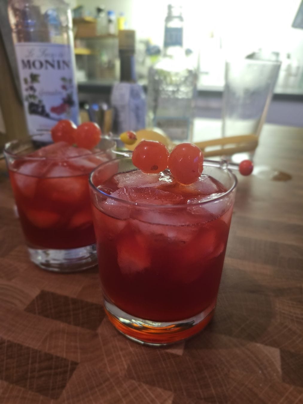 Two old fashioned glasses filled with red liquid and ice cubes. Two maraschino cherries skewered on top.