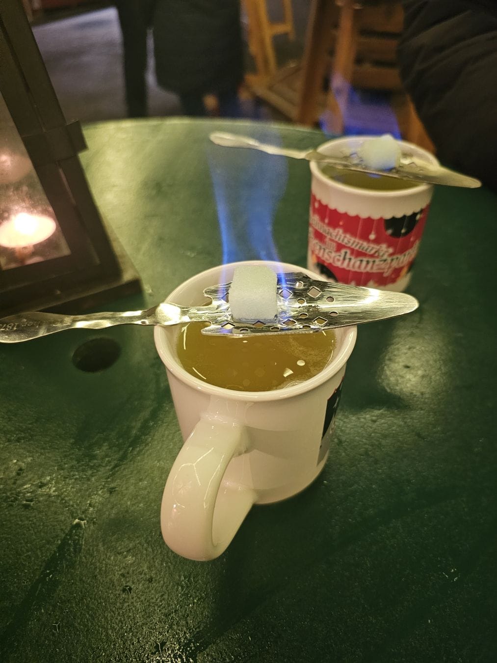 Two Absinth punch mugs on a green wooden table. Balanced across the rim, a metal grill holding a piece of burning sugar.