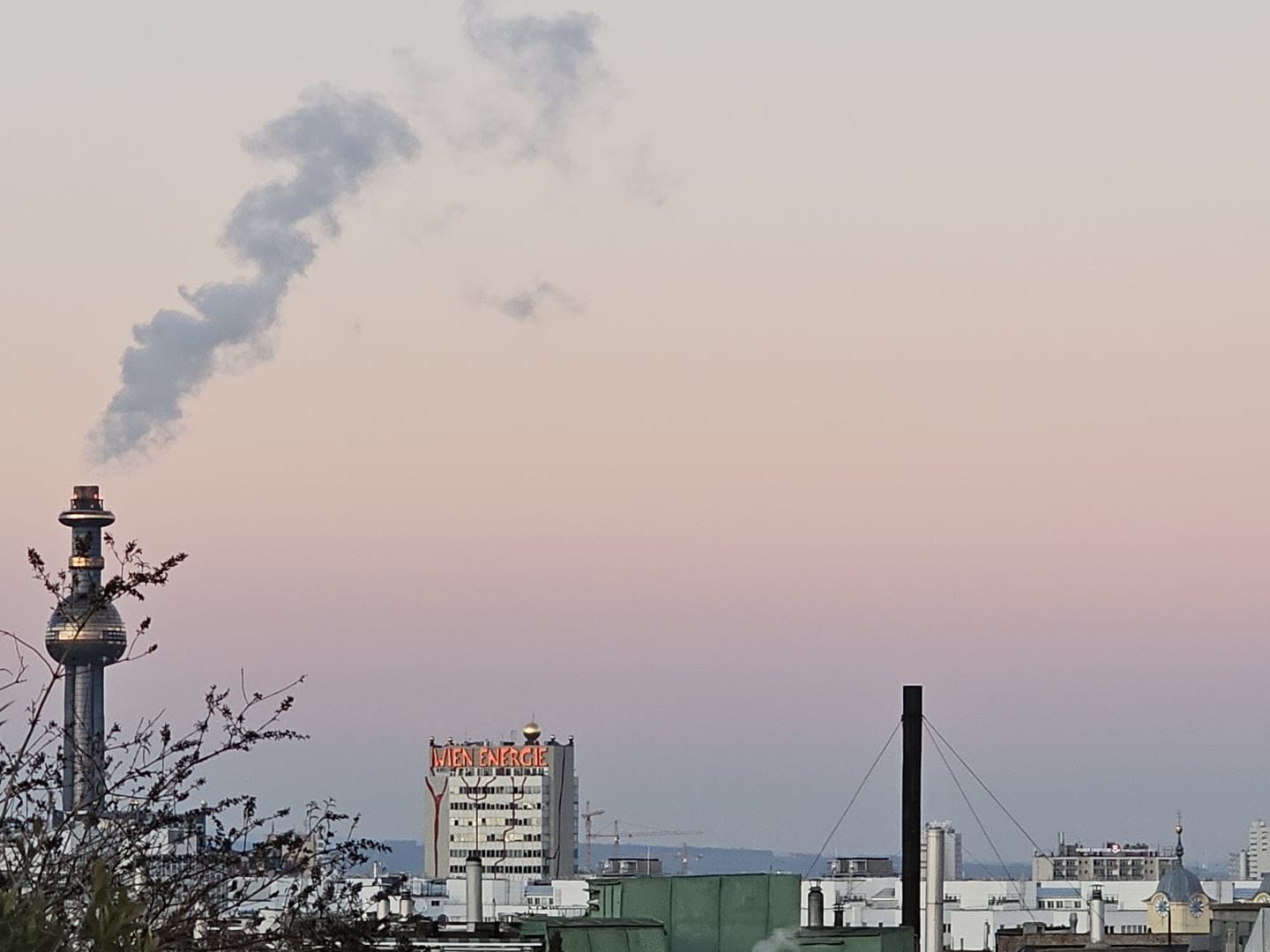Evening cityscape with industrial buildings; the Spittelau waste incineration plant chimney rises on the left, emitting a plume of smoke, with a Wien Energie office building in the center beneath a pale pink and blue sky.
