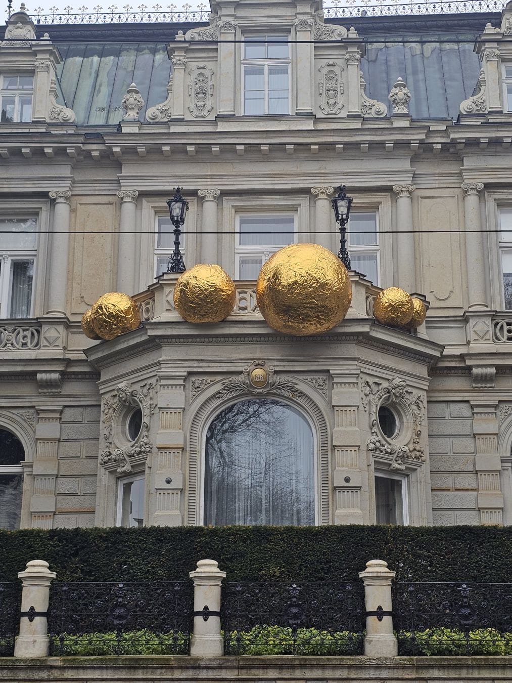 Historic stone building façade with ornate neoclassical detailing; above a central arched window sits a balcony topped by several oversized gold-colored spheres arranged symmetrically, contrasting with the pale masonry and dark roof behind.