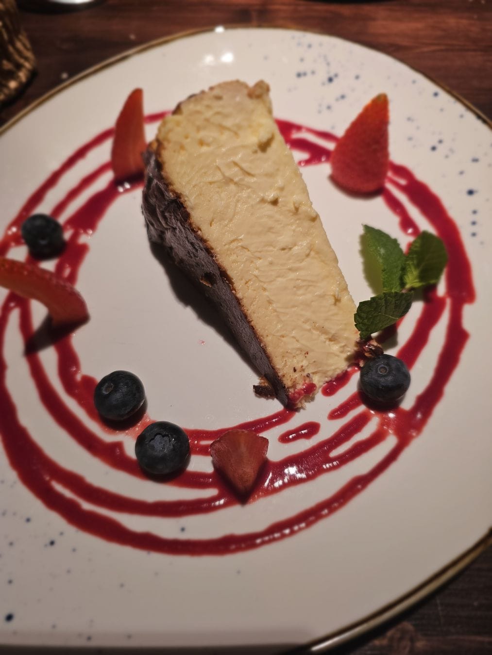 A cheese cake, on a white plate, surrounded by swirls of red fruit drizzle, an assortment of berries.