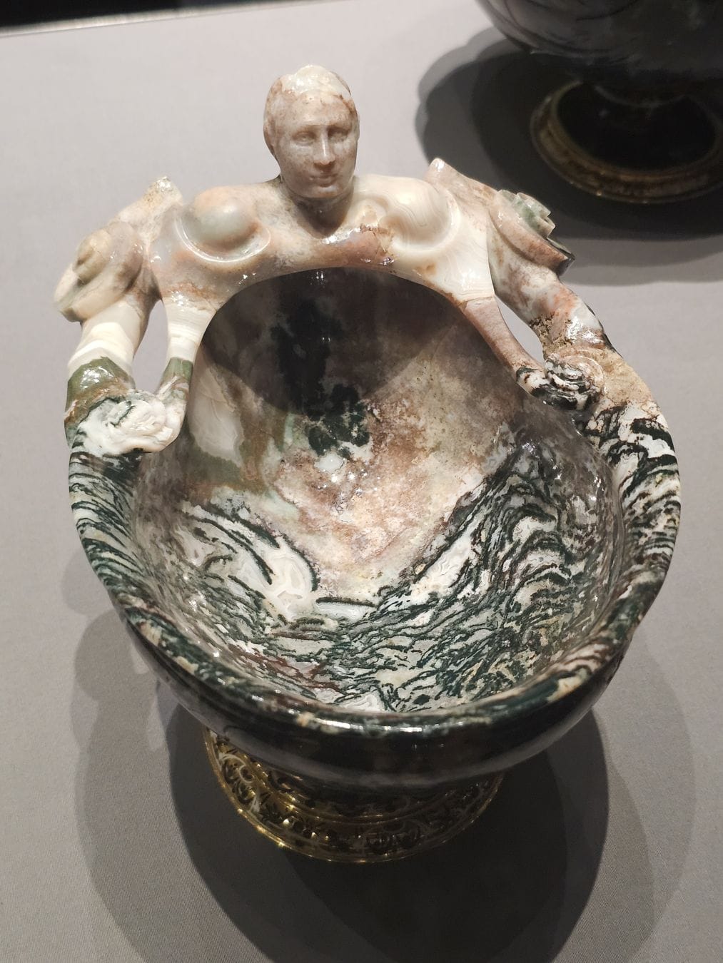 An ornate, shallow vessel carved from polished agate stone featuring swirling patterns of deep green, white, and tan. The upper rim of the bowl is sculpted into the bust of a human, the arms extending outward to form the sides of the rim. The vessel rests on a decorative gold base.