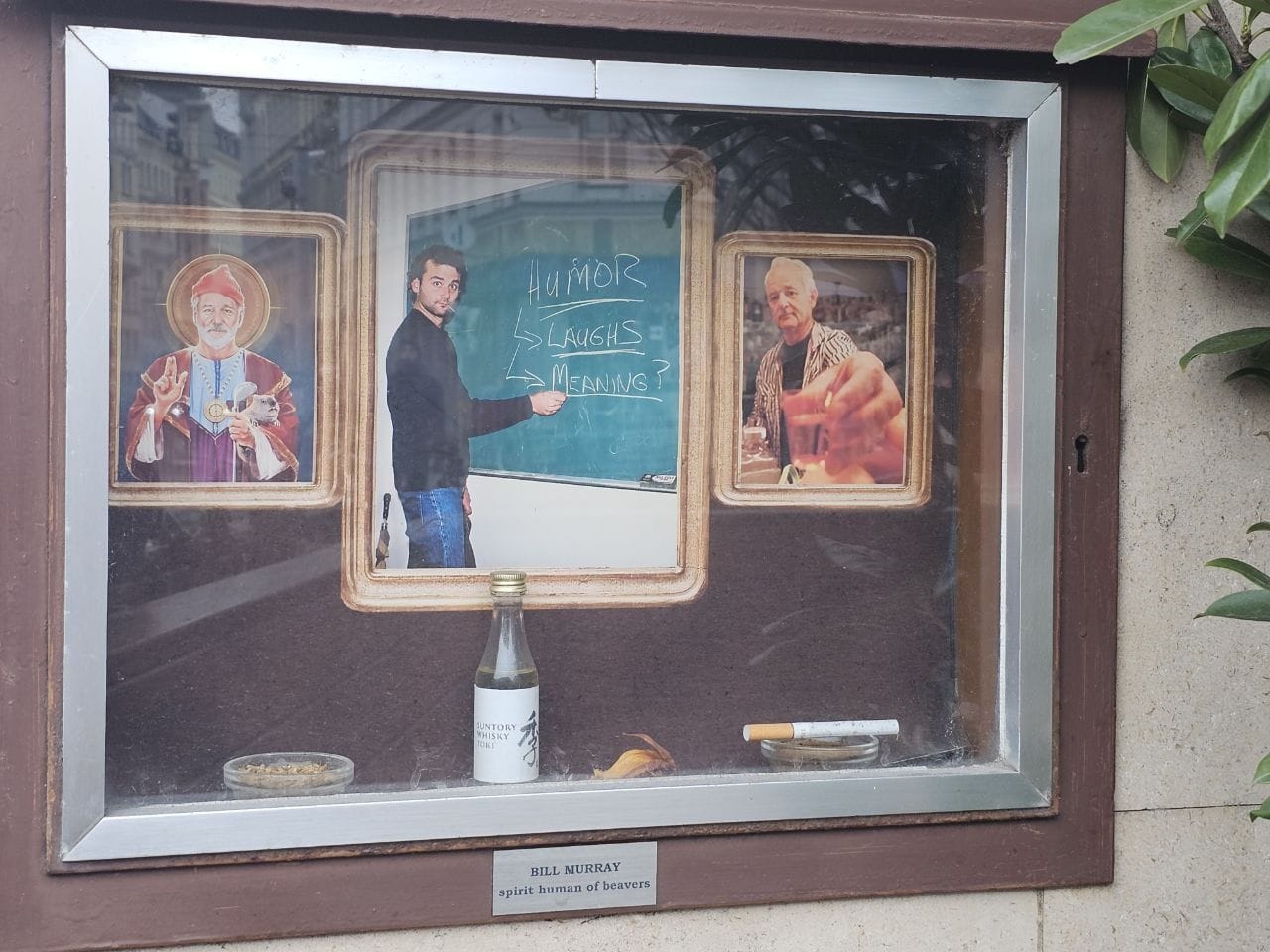 A street-level display case dedicated to Bill Murray. It features three framed photos: on the left, an icon-style painting of Bill Murray as a saint; in the center, a photo of him pointing to a chalkboard that reads 'HUMOR -> LAUGHS -> MEANING?'; on the right, a photo of him holding up a glass. Inside the case, there is also a small bottle of Suntory Whisky, a cigarette, and a small plaque that says 'BILL MURRAY spirit human of beavers'.
