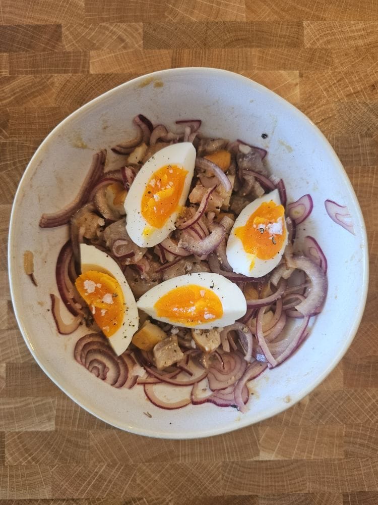 Overhead photo of a white bowl on a wooden butcher-block counter, with thinly sliced red onion, pieces of marinated sherry herring, and Schillerlocken (smoked dogfish belly strips), plus four wedges of soft-boiled egg with jammy orange yolks, finished with cracked black pepper and coarse salt.