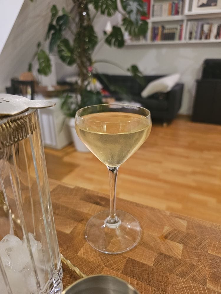 A clear, pale-yellow cocktail in a Nick & Nora glass sits on a wooden butcher-block counter. In the foreground, a crystal mixing glass with ice and a cocktail strainer is visible. The background shows a cozy living room with a dark couch, a large Monstera plant, and a white bookshelf filled with books.