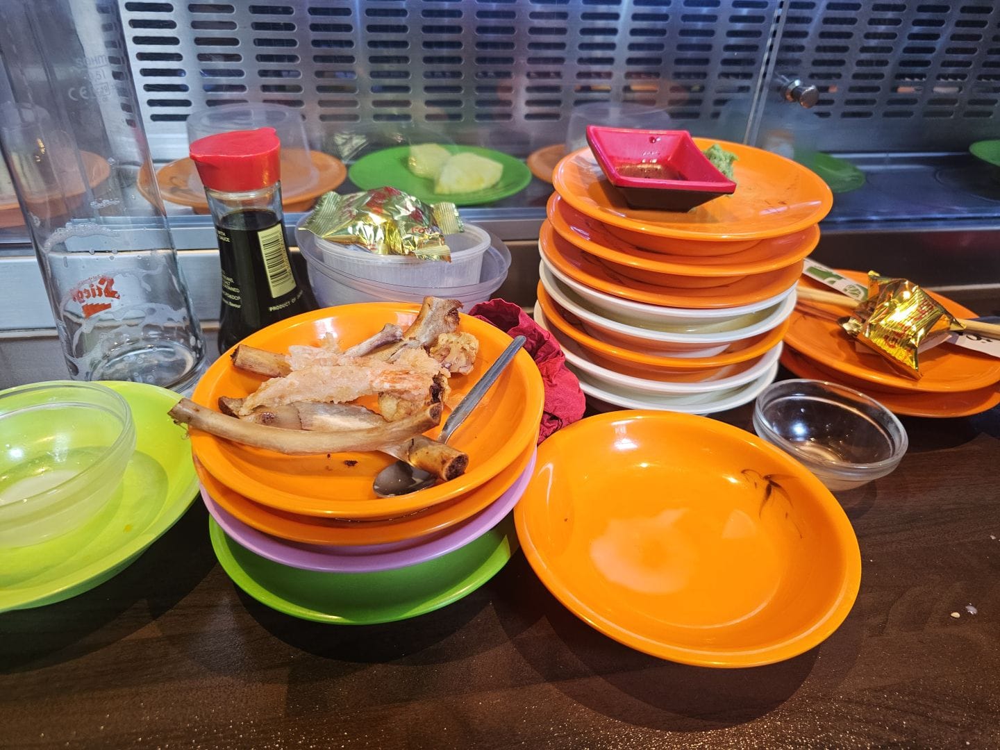 The aftermath of a conveyor-belt sushi feast: tall stacks of empty color-coded plates beside a sushi belt. One plate holds picked-clean rib bones. A bottle of soy sauce, an empty beer glass, and scattered remnants complete the scene of a hearty meal.
