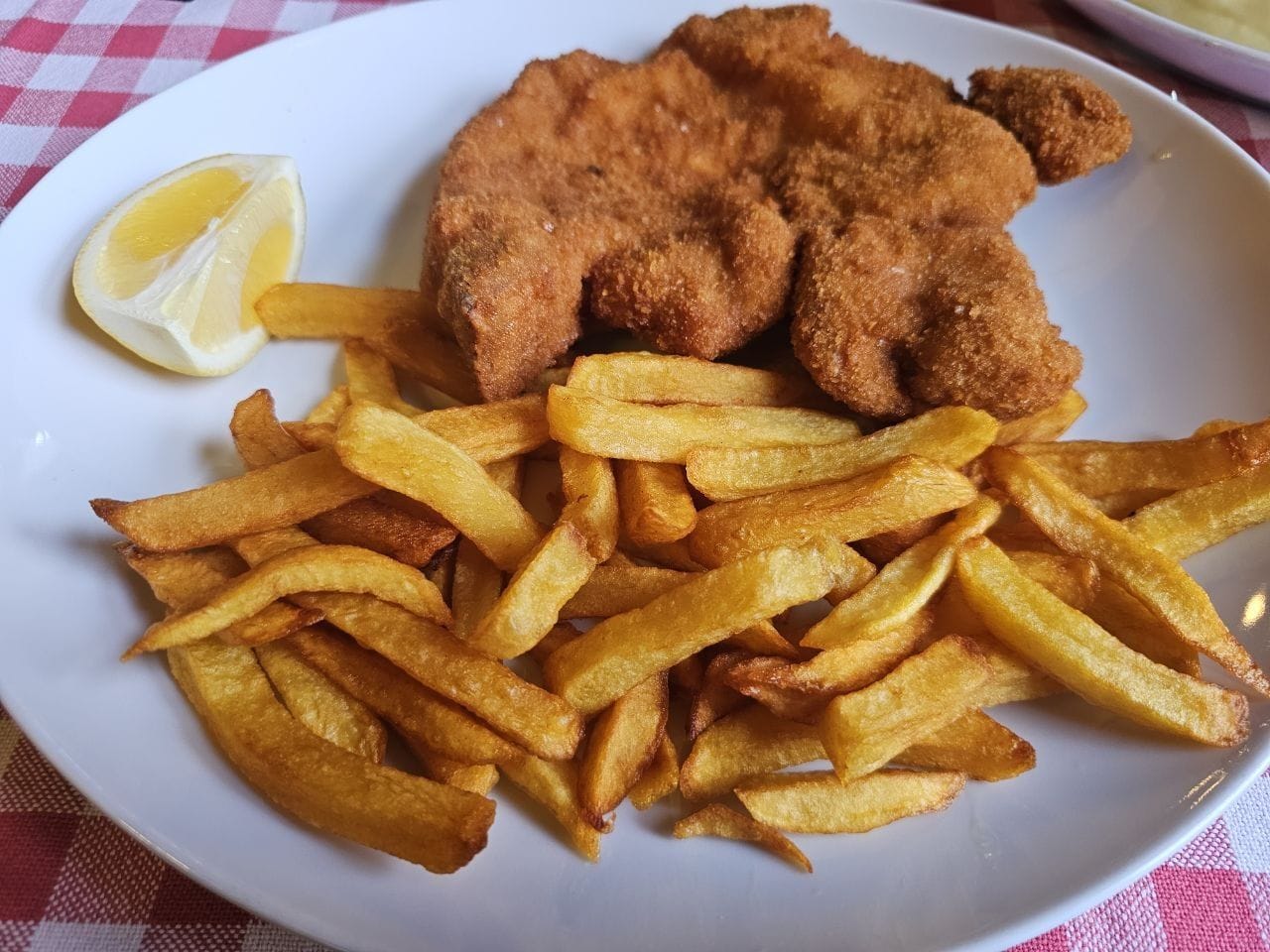 Fried pork cutlet served with thick-cut fries and a lemon wedge on a white plate, placed on a red and white checkered tablecloth.