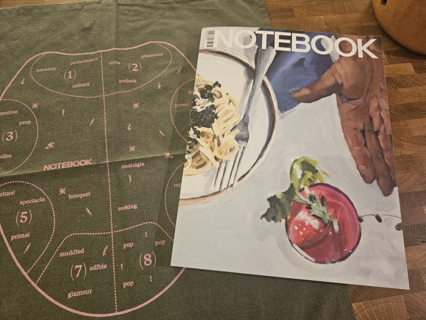 A napkin and a NOTEBOOK publication on a wooden surface. The left item is an olive green napkin with pink text showing a circular diagram divided into 8 sections with words like 'uncanny,' 'performance,' 'blobs,' 'phantoms,' 'nostalgia,' 'pop,' 'glamour,' and 'edible.' The right item is a NOTEBOOK publication featuring a still life painting of a white plate with pasta, sausage, and a strawberry with chocolate sauce.