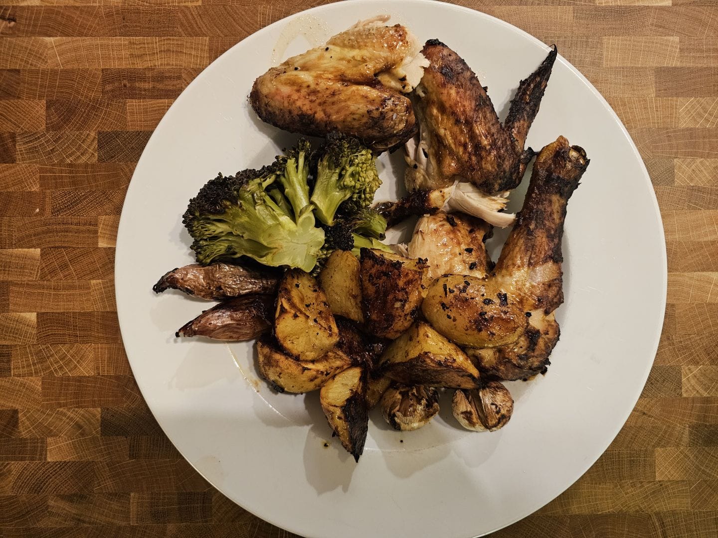 Roast chicken pieces with potatoes, broccoli, shallots and garlic on a white plate on a butcher's block surface.