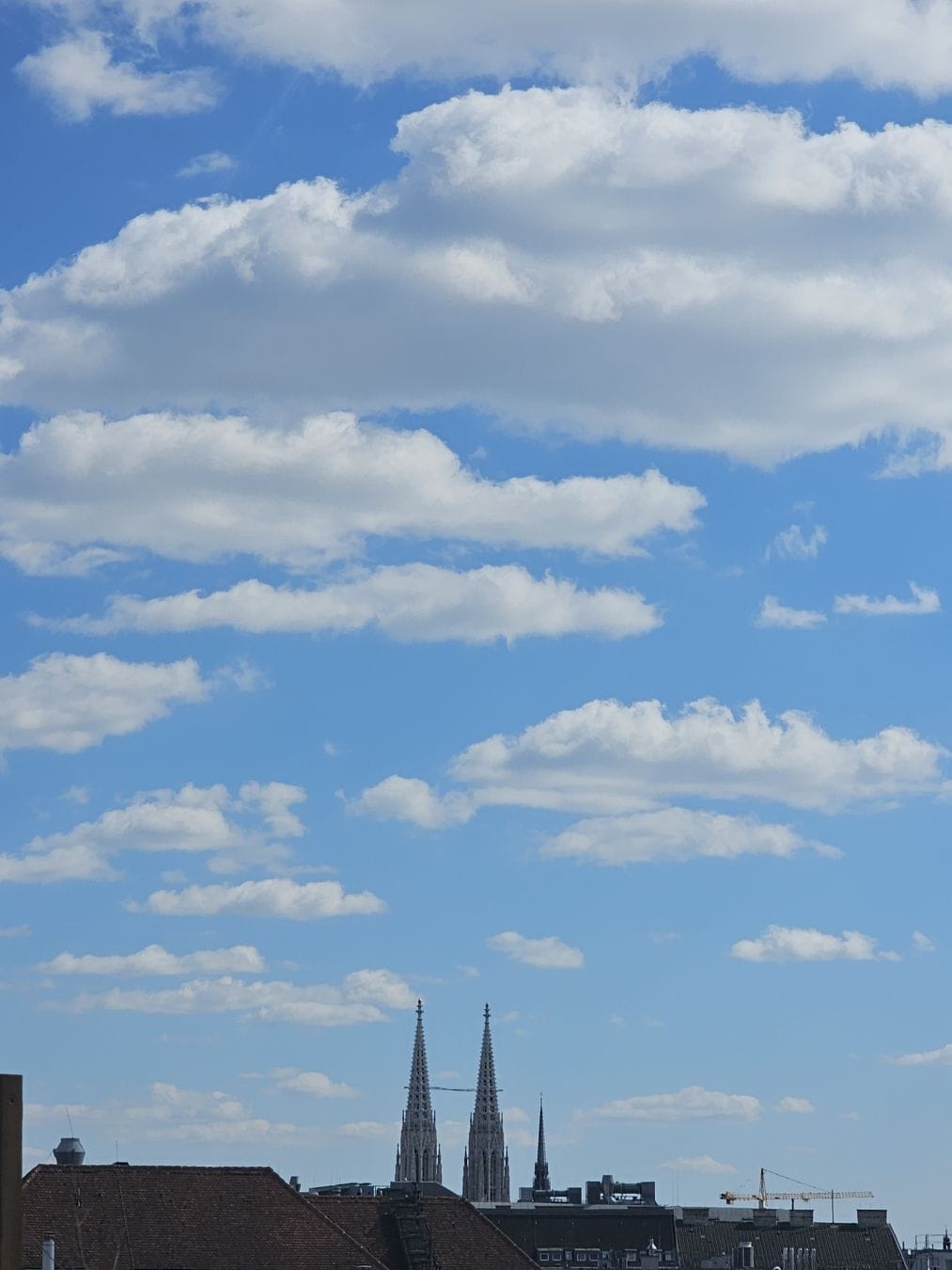 White clouds against a blue sky, Votiv church and various other tops of houses of Vienna visible in the bottom.