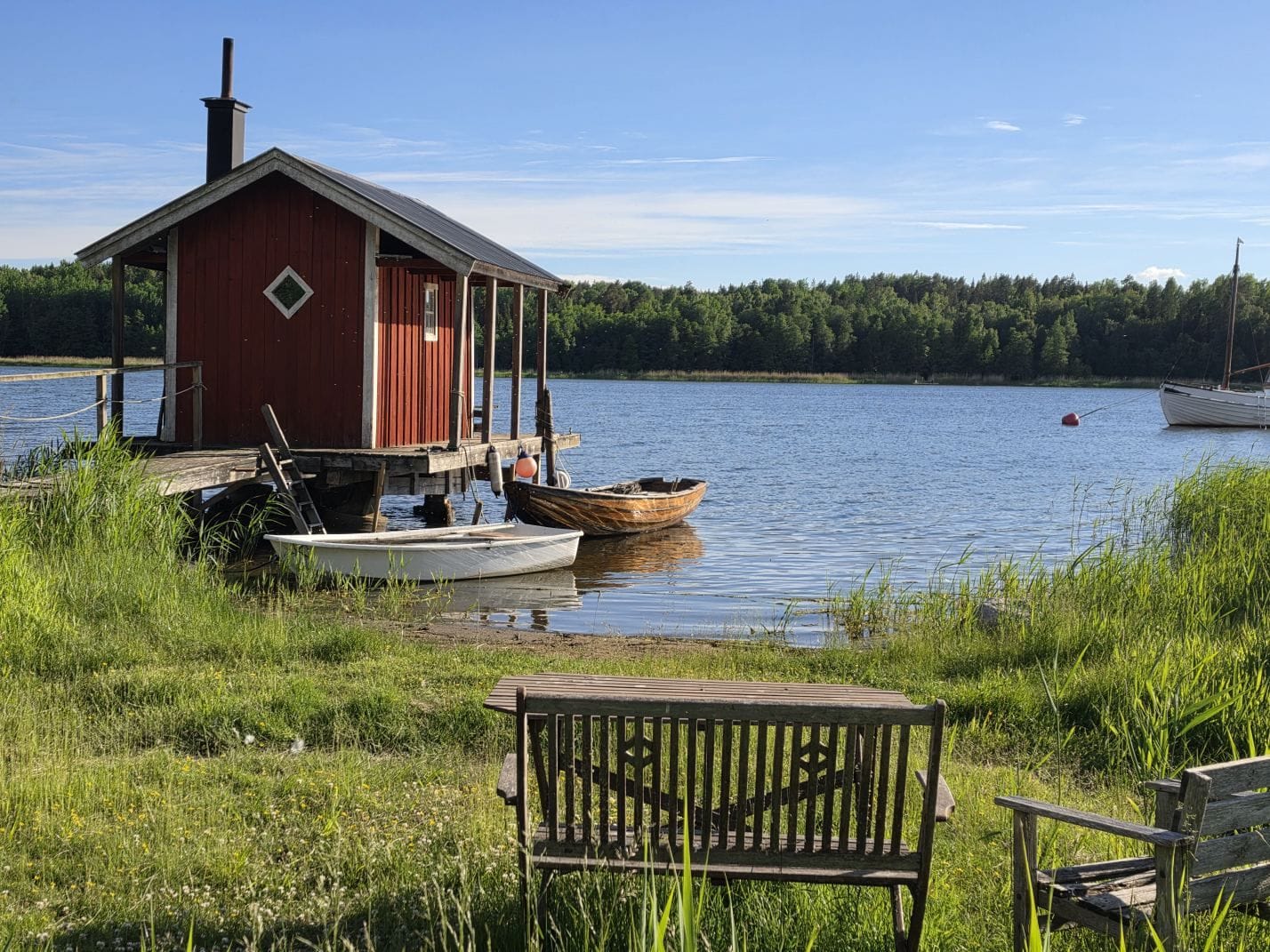 A red boathouse, small boats and wooden benches beside calm water under a blue sky.