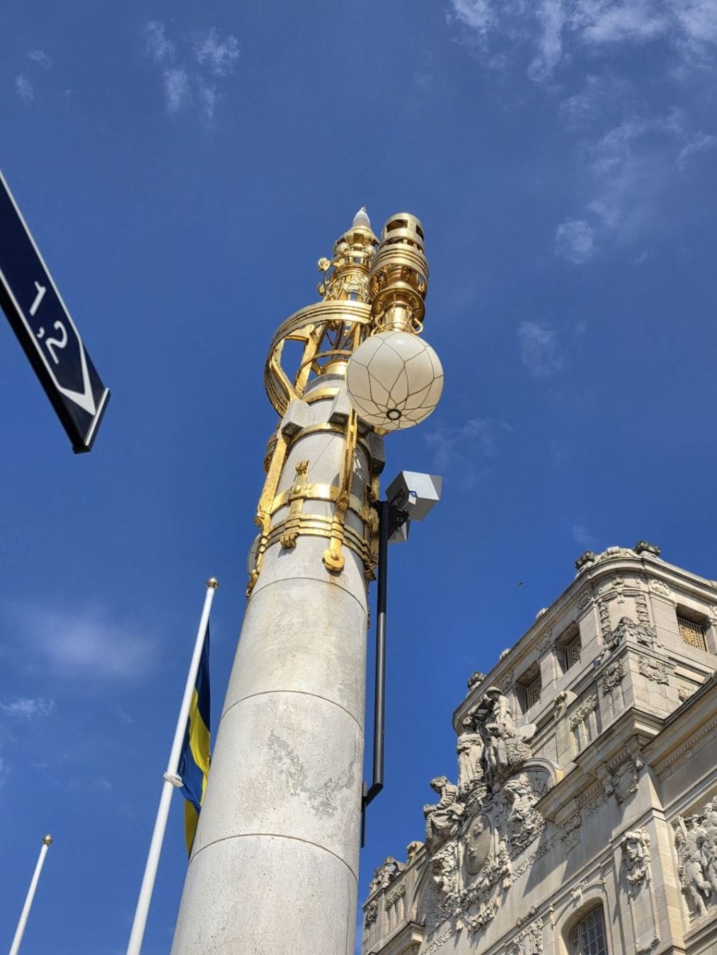A gilded streetlamp rises beside an ornate stone building and Swedish flags under a bright blue sky.