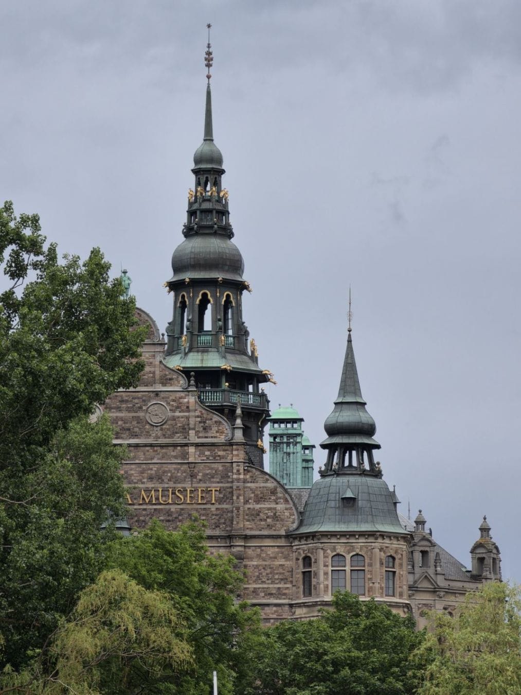 Nordiska museet’s brick facade and green copper towers rise behind trees under a grey sky.