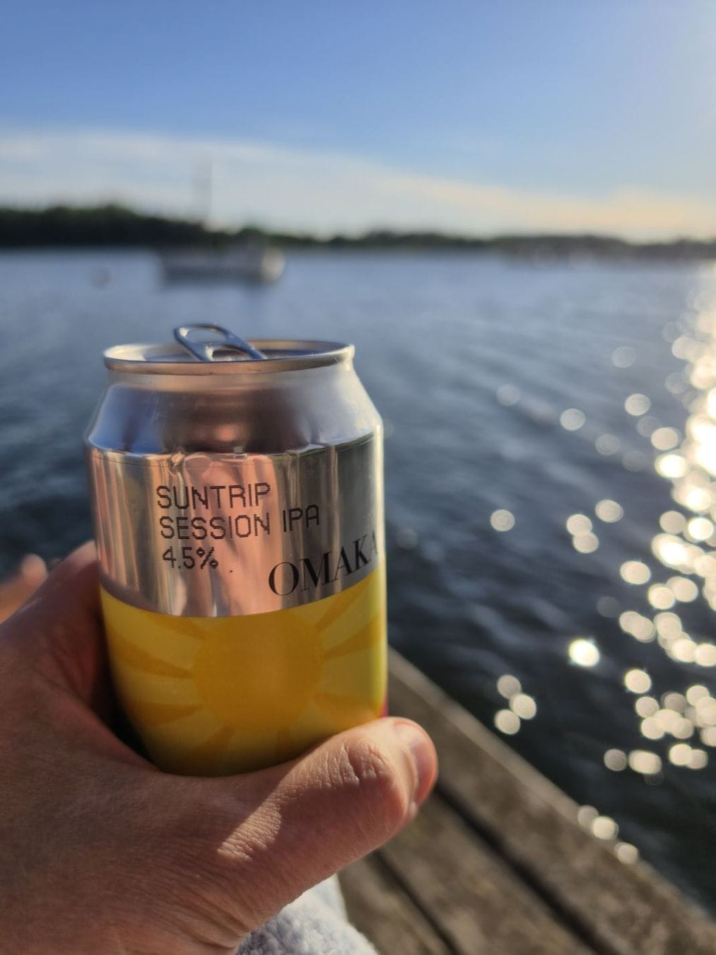 A hand holding a yellow beer can beside sunlit water with boats blurred in the background.