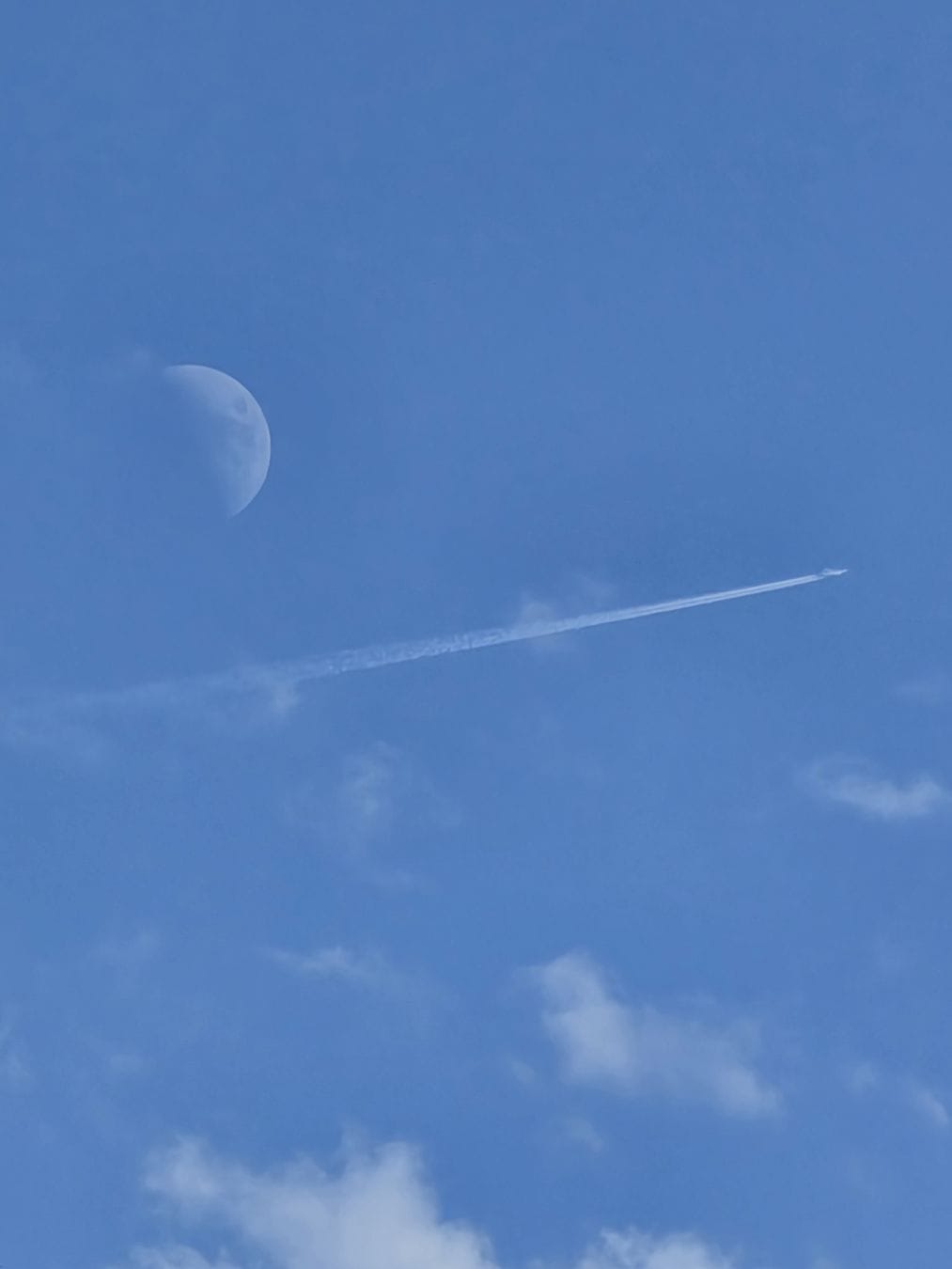 Blue sky, to the left a waxing moon, below the contrails of a plane leaving towards to the right side of the picture. Below a few scattered, airy clouds.
