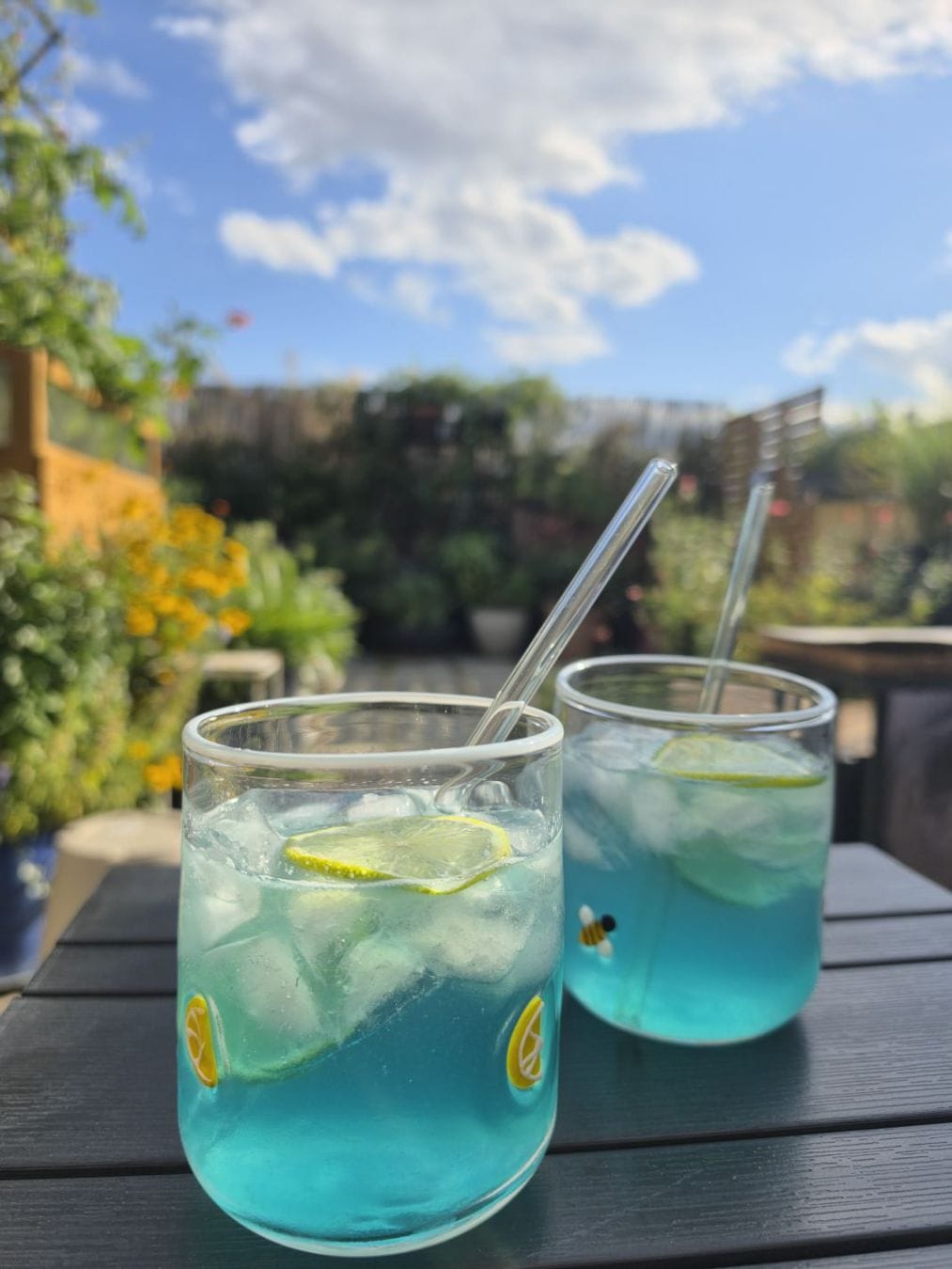 Two bright turquoise iced drinks with lime slices and glass straws on a sunny garden table beneath a blue, cloud-dappled sky.