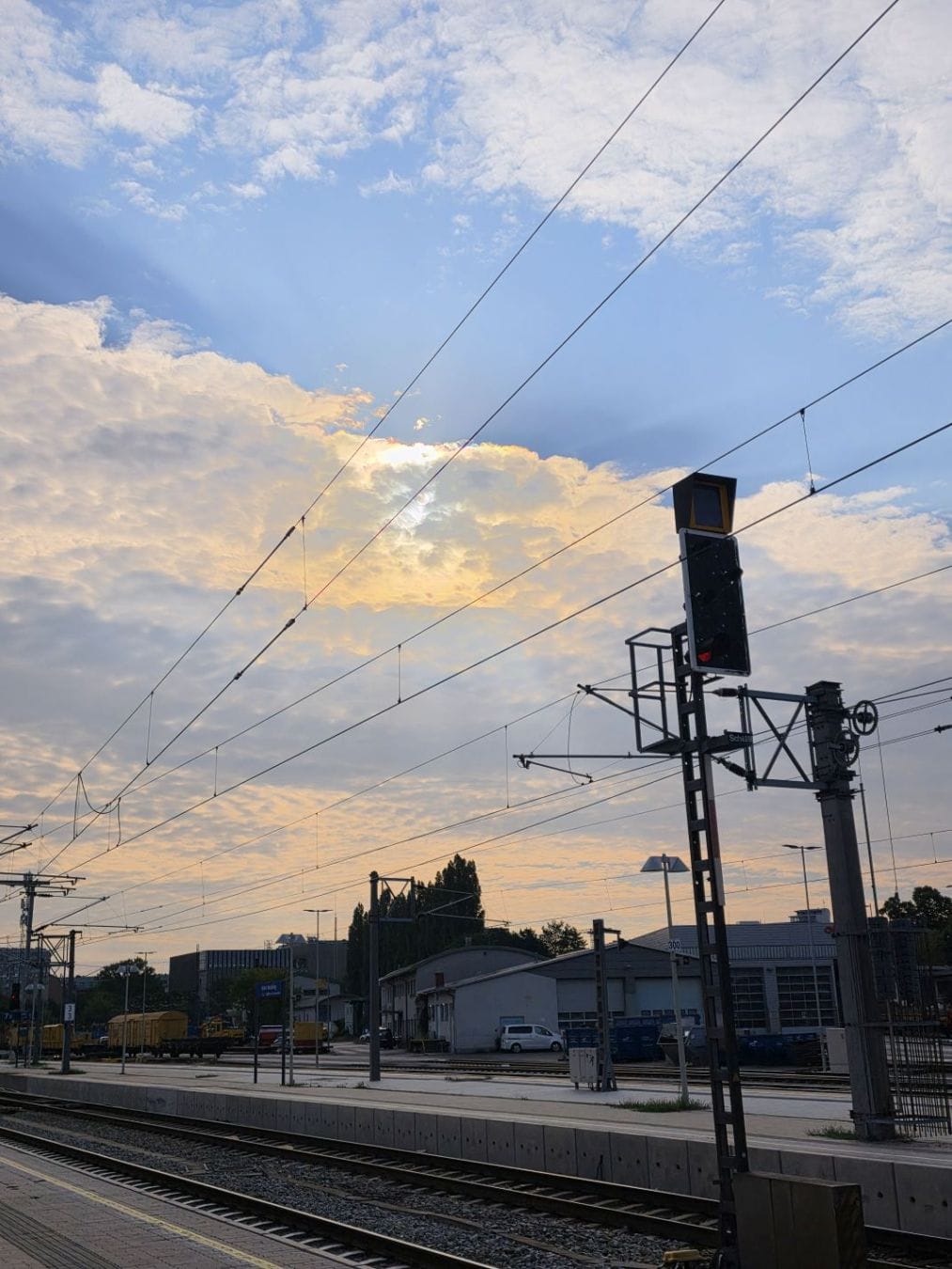 Railway tracks and overhead power lines beneath sunlit clouds, industrial buildings in the background.