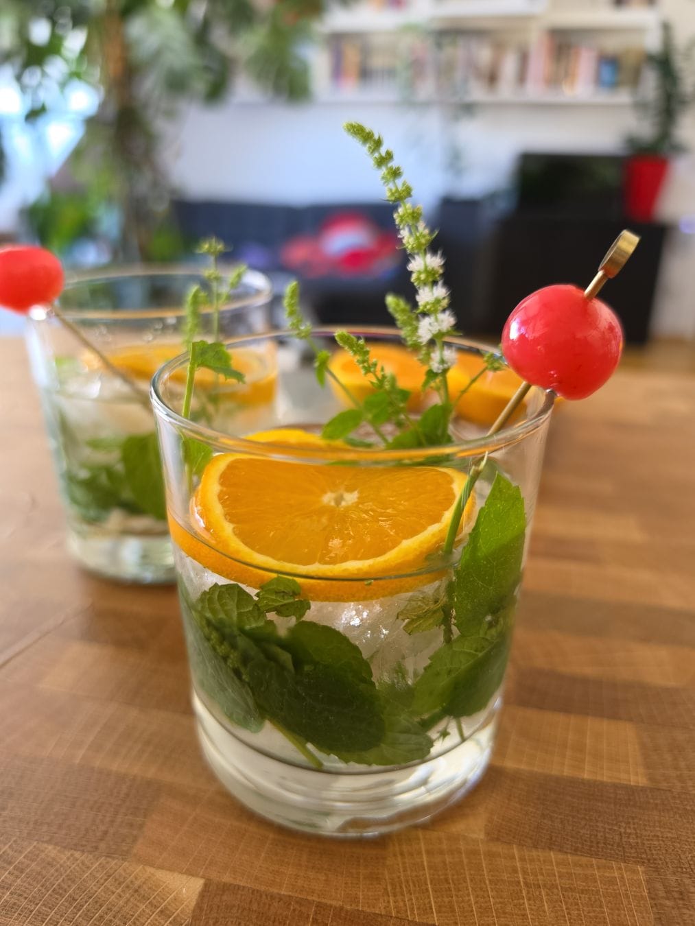 A clear liquid in an old fashioned glass with muddled mint and ice, also a slice of orange, a mint sprig with blossoms and a cocktail stick with cherry. In the background, the same thing. Everything on a wodden surface.