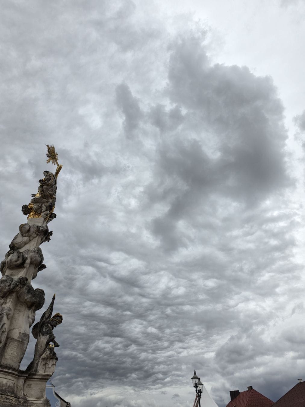 An ornate stone column with gilded details rises beside rooftops beneath a vast sky of textured grey clouds.