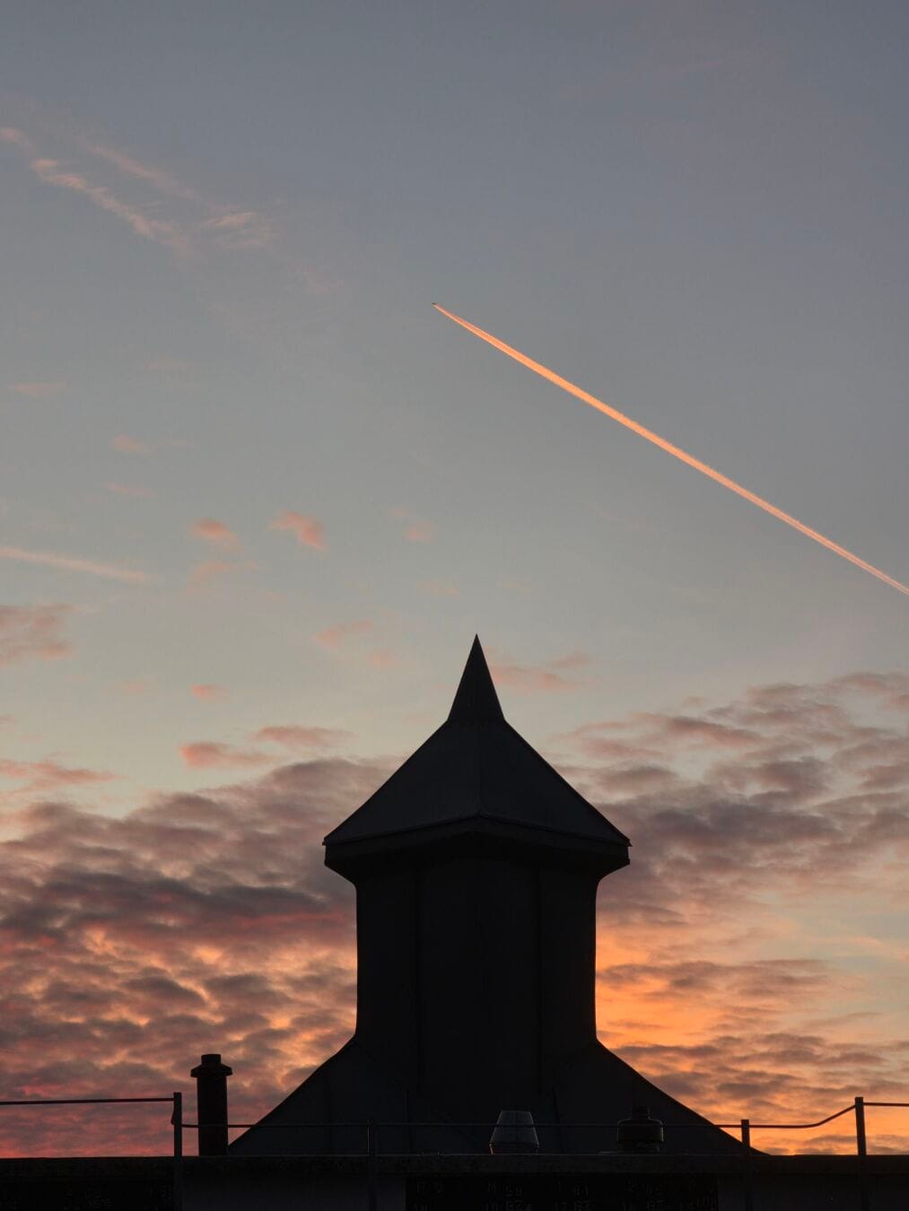 A pointed rooftop tower silhouetted against orange and pink clouds, with a plane trailing a glowing orange contrail overhead.